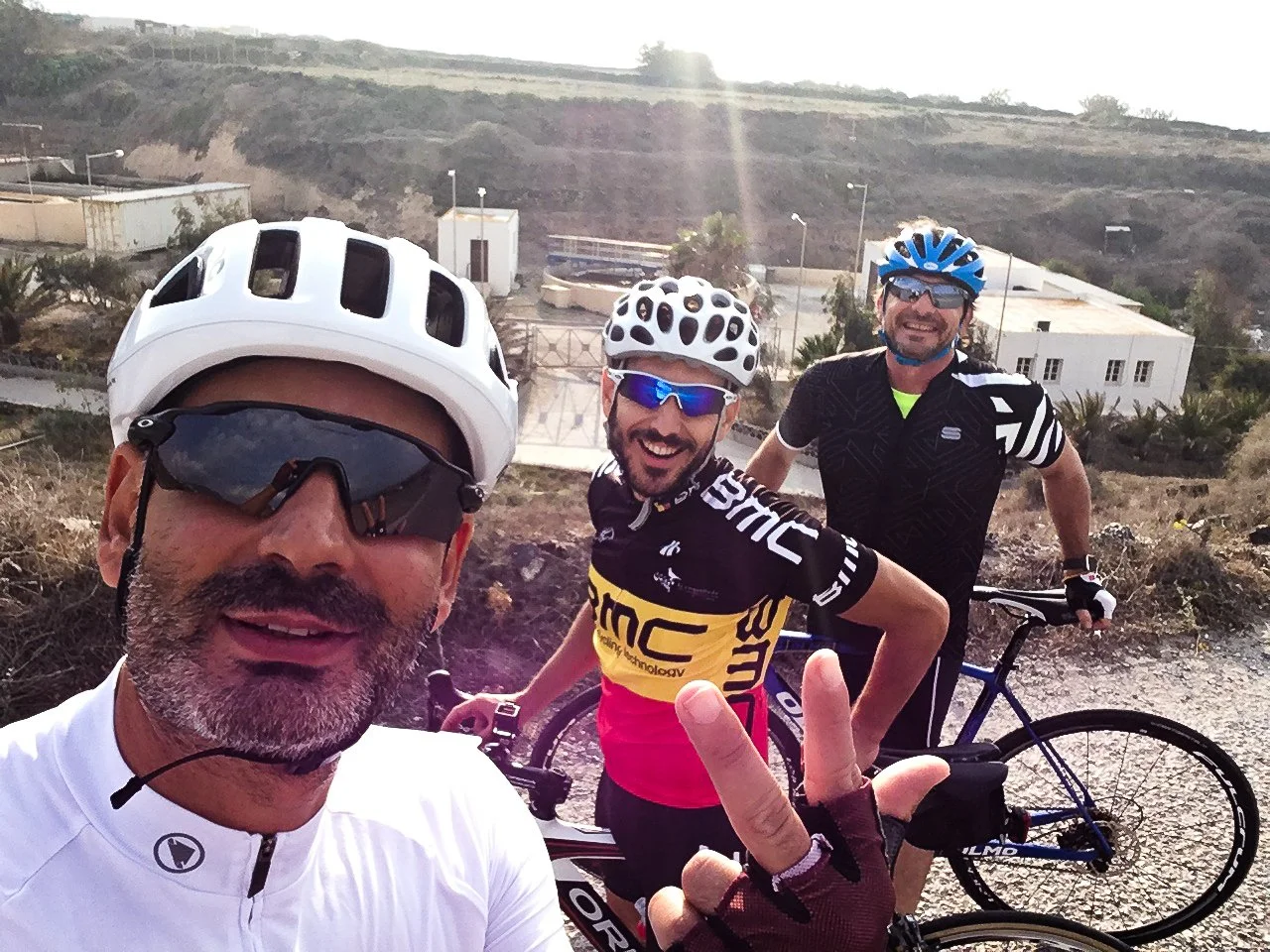 Three male cyclists smiling and taking a selfie outdoors with a bicycle in the background. They are wearing helmets and cycling gear, with one showing a peace sign. The background includes buildings and a hillside.