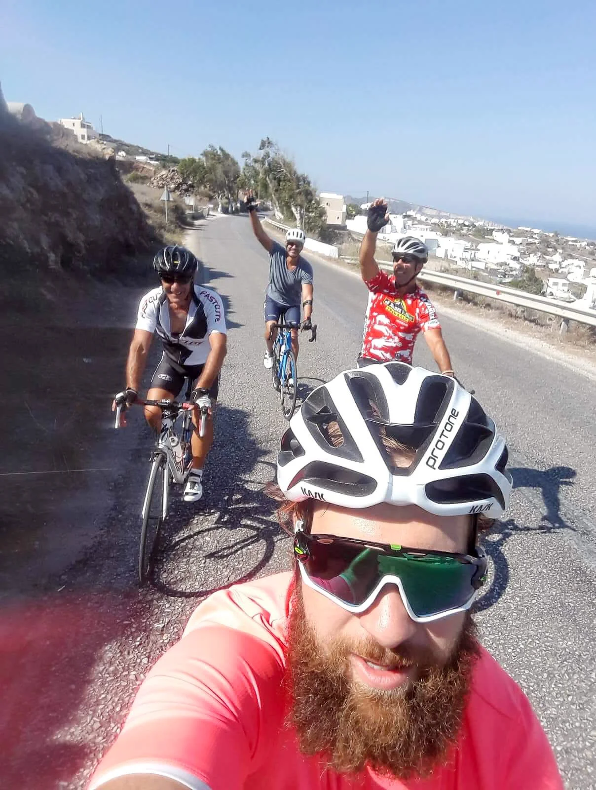A group of four cyclists taking a selfie on a mountain road, with two waving and smiling, in a sunny, hilly landscape with houses in the background.