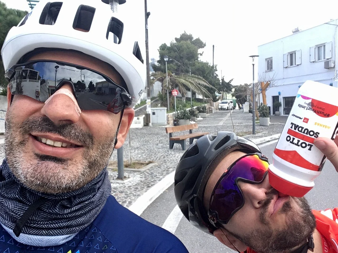 Two cyclists in helmets and sports sunglasses, one smiling at the camera, the other drinking from a sports drink cup, standing in a paved outdoor area with benches, trees, and buildings in the background.