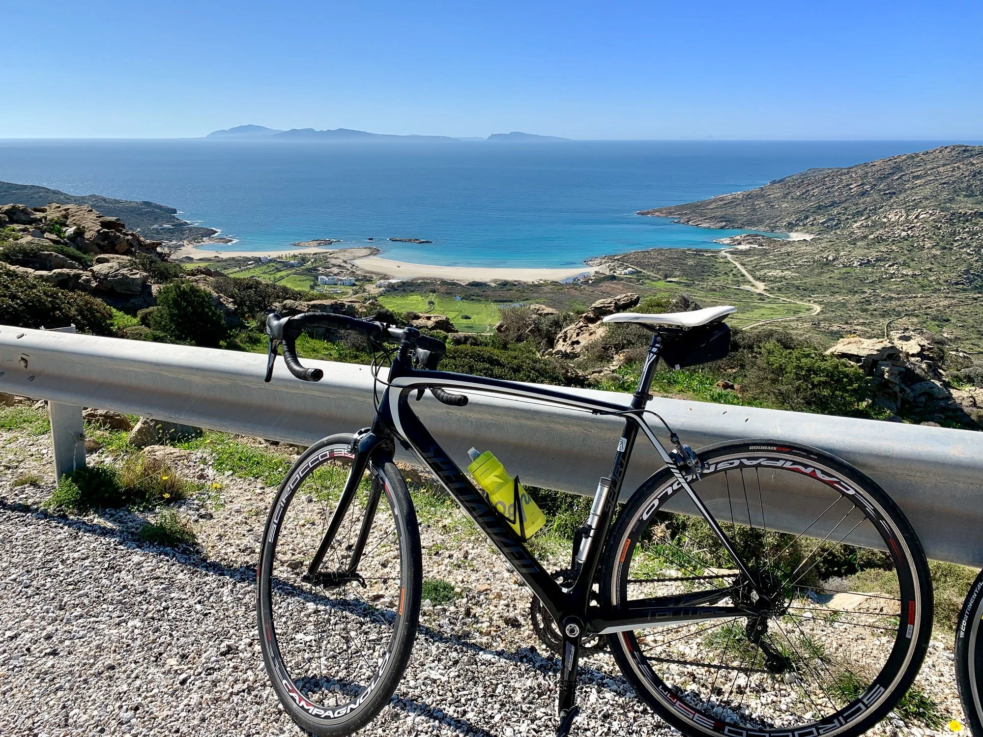 A black and white road bicycle leaning against a white guardrail overlooking a coastal landscape with mountains, a beach, and the ocean in the background.