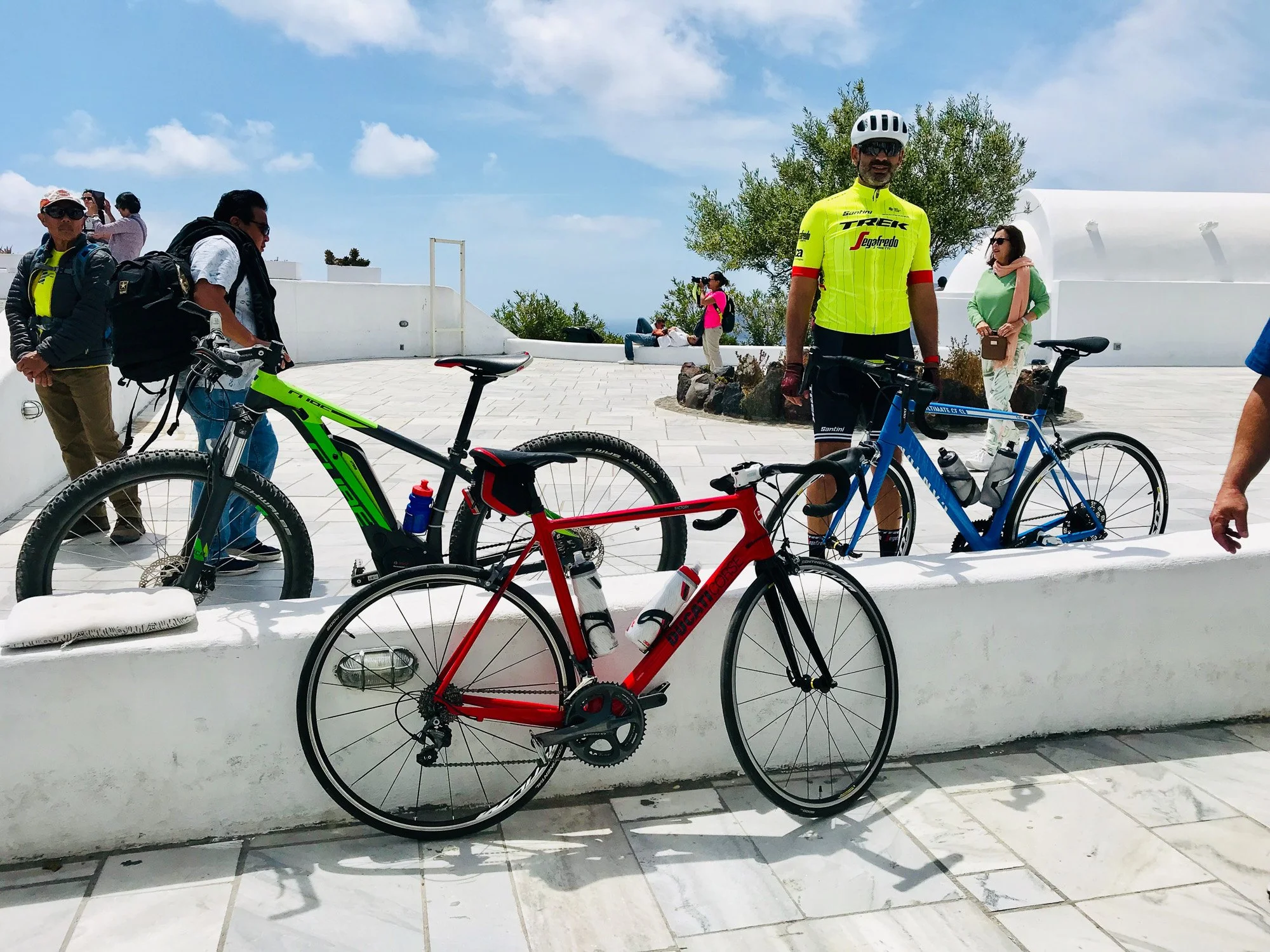 Group of cyclists and tourists in a sunny outdoor location with white architecture and blue sky, some standing near bikes and others sitting or walking.