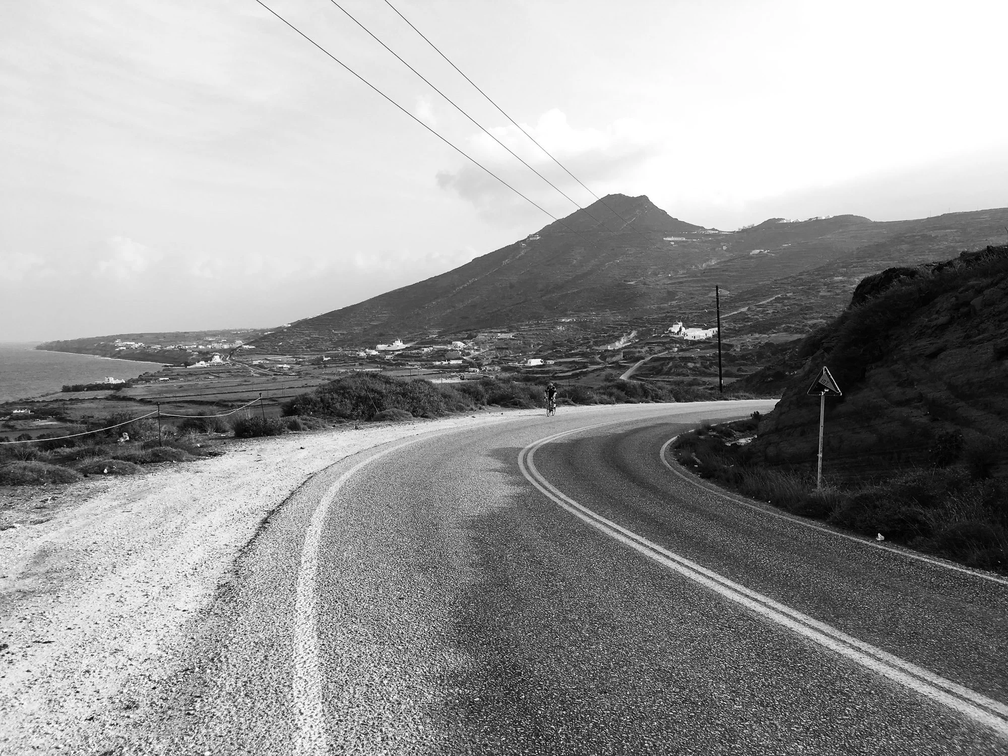 A black and white photo of a winding coastal road with a cyclist, mountainous landscape in the background, utility poles, and a warning sign on the roadside.