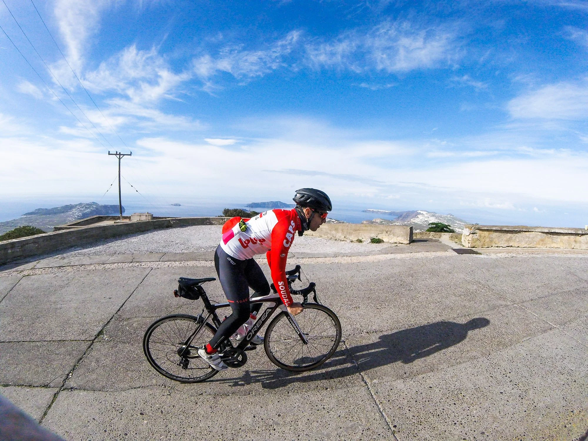 Cyclist riding on a mountain road with a scenic view of the coastline, hills, and blue sky with clouds.