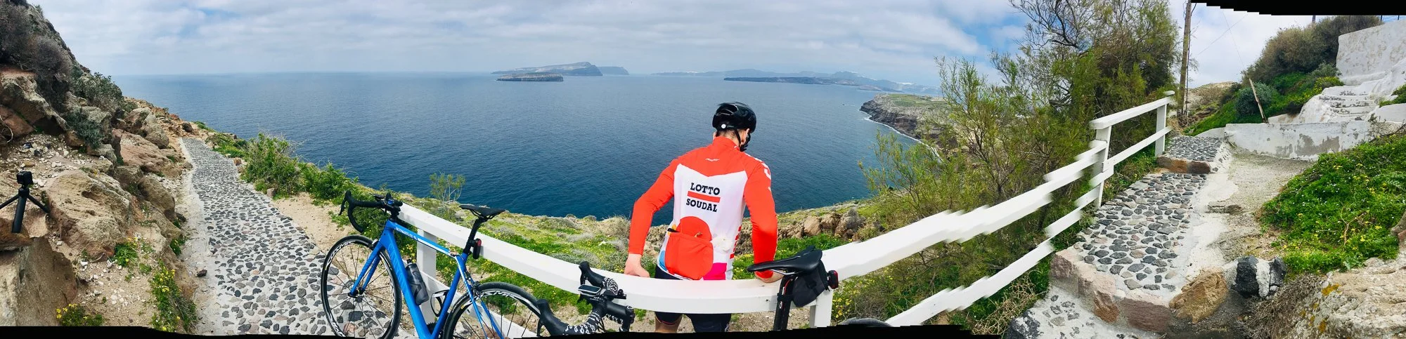 Cyclist standing on a cobblestone path overlooking the ocean, with bicycles nearby and a white fence along the trail.