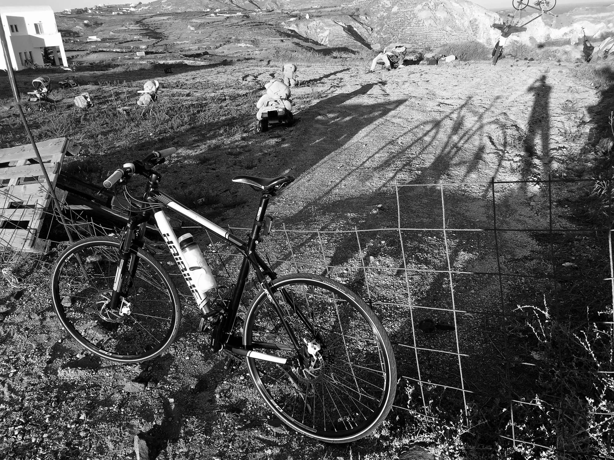 A mountain bike resting against a wire fence in a rural outdoor area with a trail, some construction materials, and a few workers in the background.
