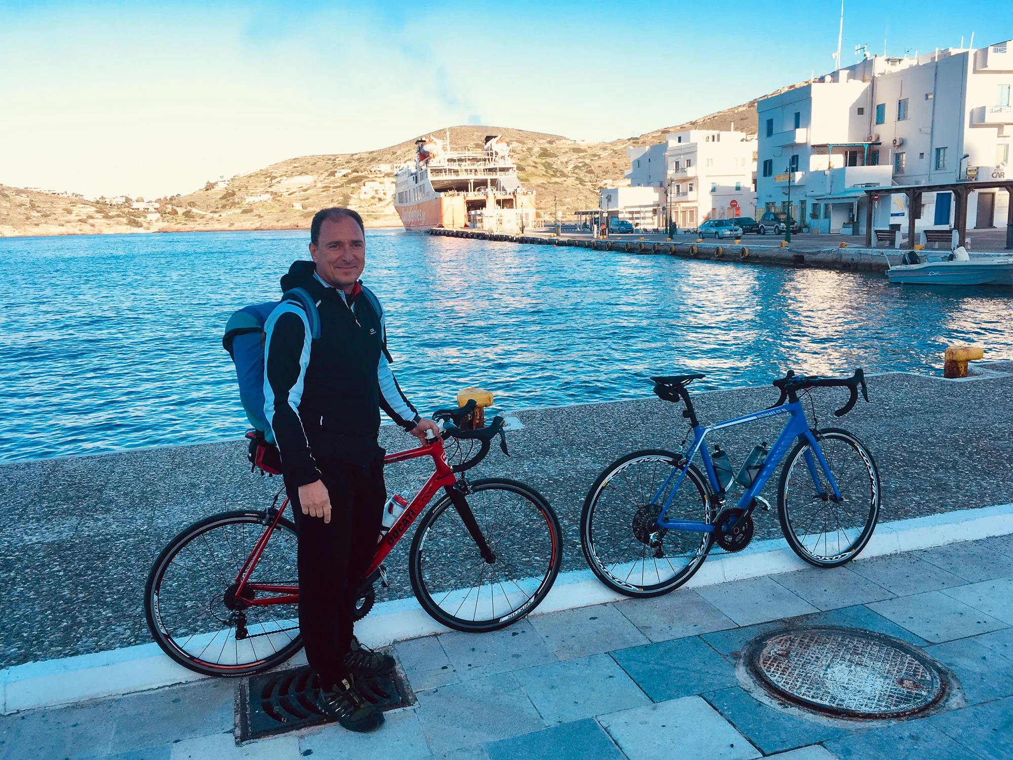 A man standing beside two bicycles on a waterfront with a boat and white buildings in the background.