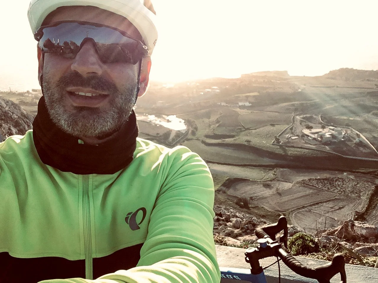 A man takes a selfie while mountain biking in a hilly, rural area near the coast during sunset. He is wearing sunglasses, a helmet, a neon green cycling jacket, and a neck gaiter. There is a mountain bike handlebar visible in the foreground. The back