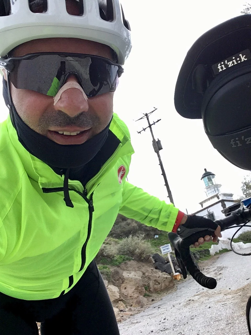 A man wearing a white helmet and black sunglasses rides a bicycle near a lighthouse and a power pole on a cloudy day.