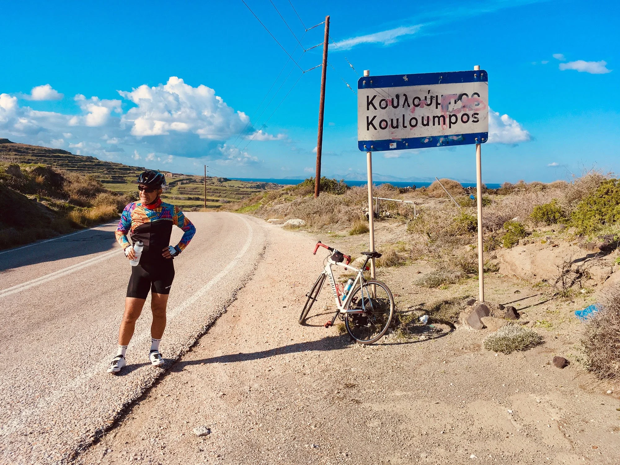 A cyclist in colorful gear holding a water bottle stands next to a white bicycle on a rural road, with a sign that reads 'Kouloumpos' in Greek and English, under a bright blue sky with scattered clouds, surrounded by arid terrain and distant hills.
