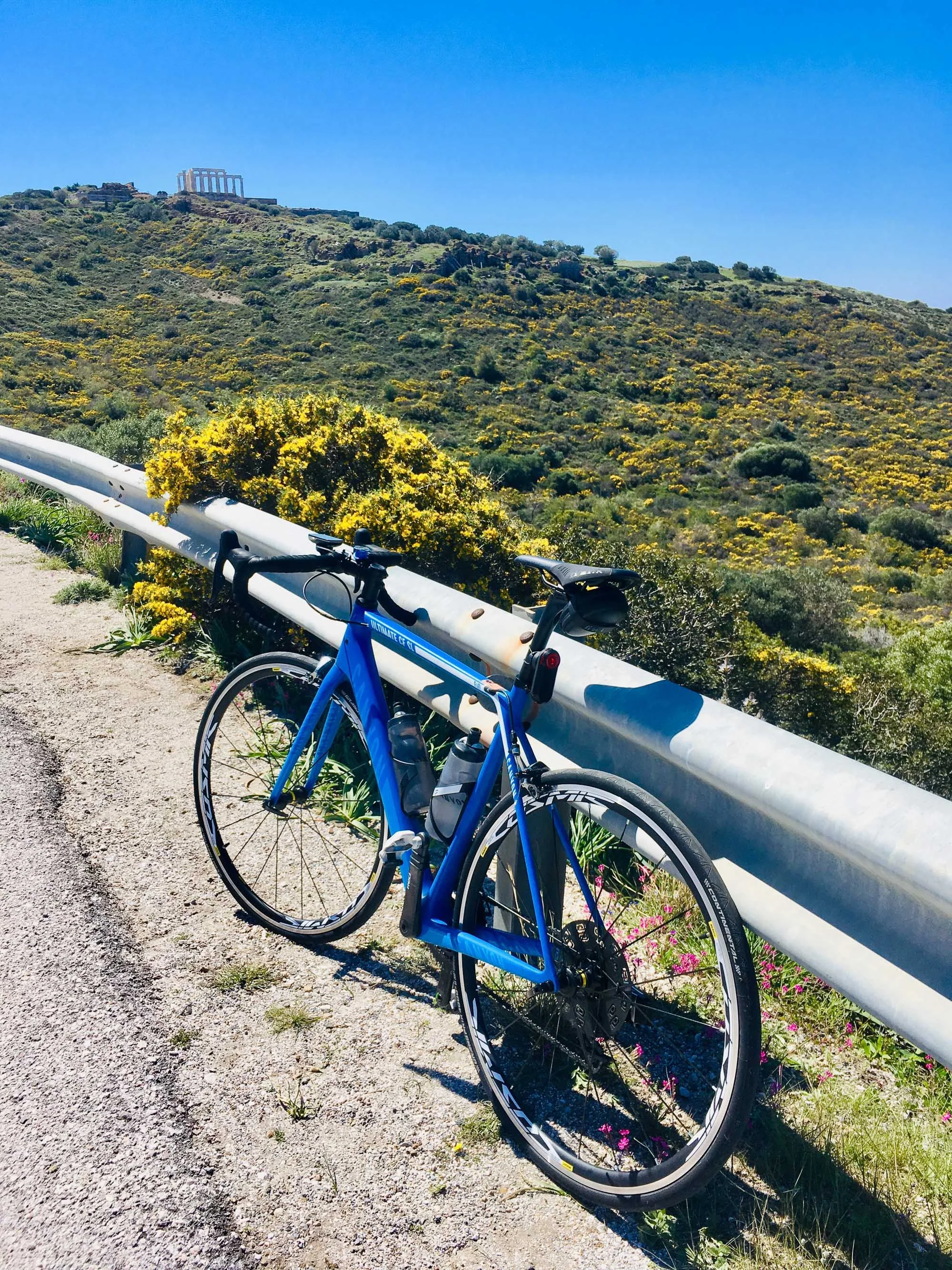 Blue road bicycle leaning against a guardrail on a hillside road with yellow flowering shrubs, green hills, and the Parthenon ruins on top of a hill in the background under a clear blue sky.