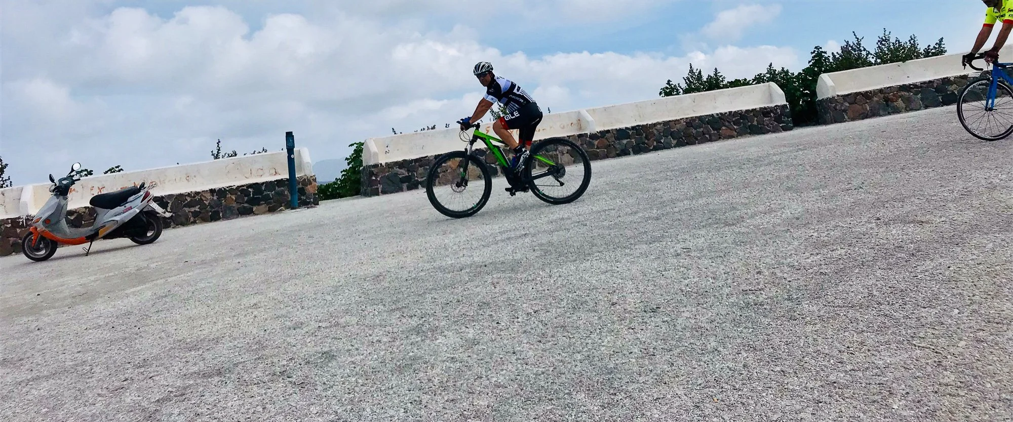 Two cyclists riding on a gravel road with stone barriers and a scooter parked to the side, under a cloudy sky, with trees in the background.