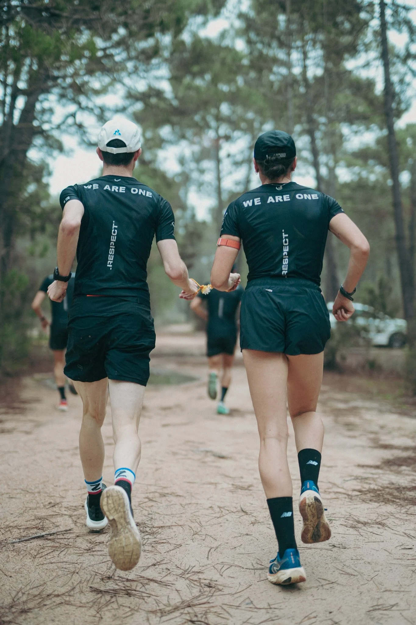 Deux coureurs en tenue de sport traverse un sentier boisé en forêt, l'un porte une casquette blanche et l'autre un bonnet noir, avec le message « WE ARE ONE » imprimé sur leurs t-shirts.