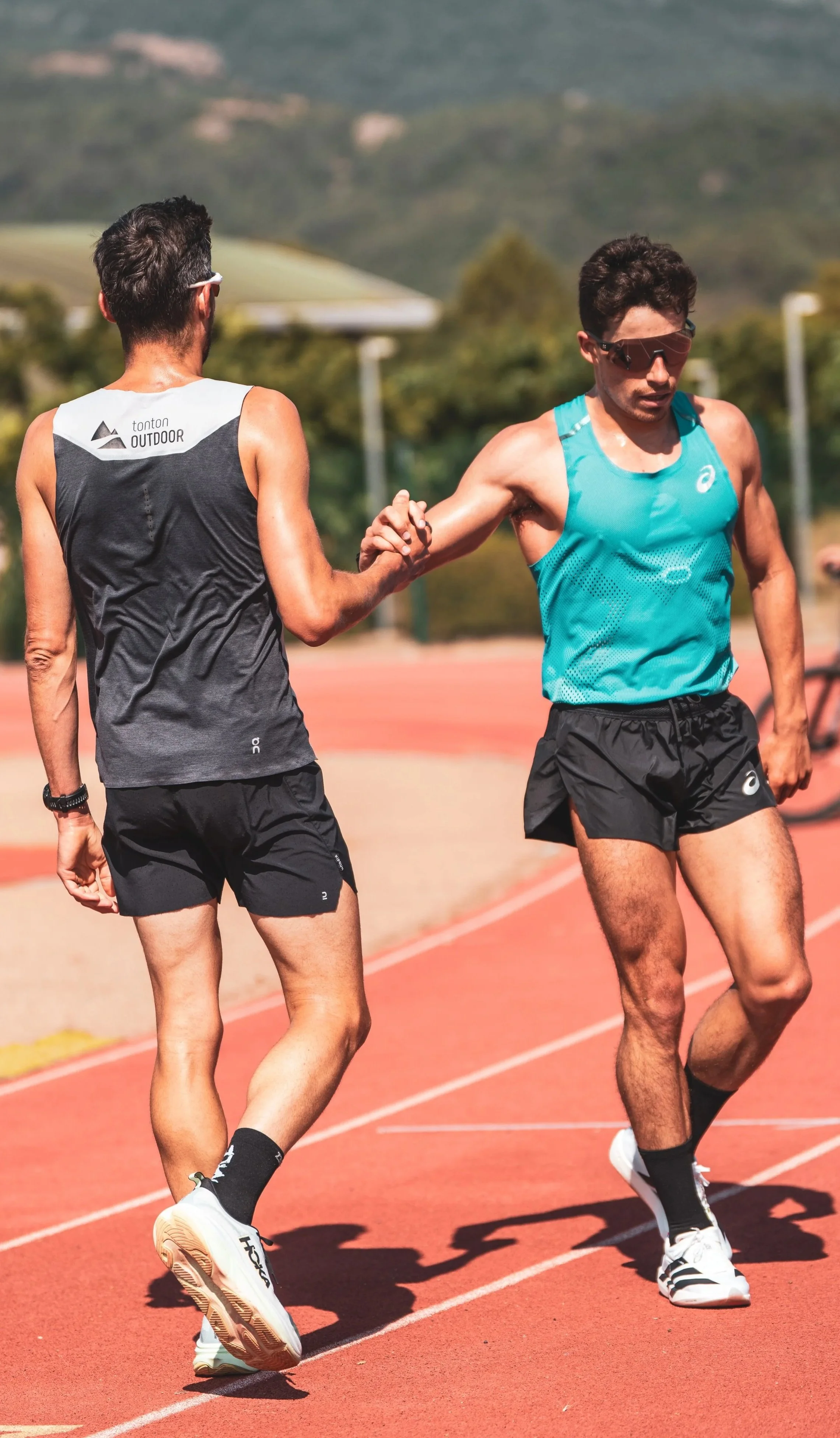 Deux hommes coureurs de marathon se donnent la main sur une piste d'athlétisme en plein air avec des montagnes en arrière-plan, portant des tenues de sport et des lunettes de soleil.
