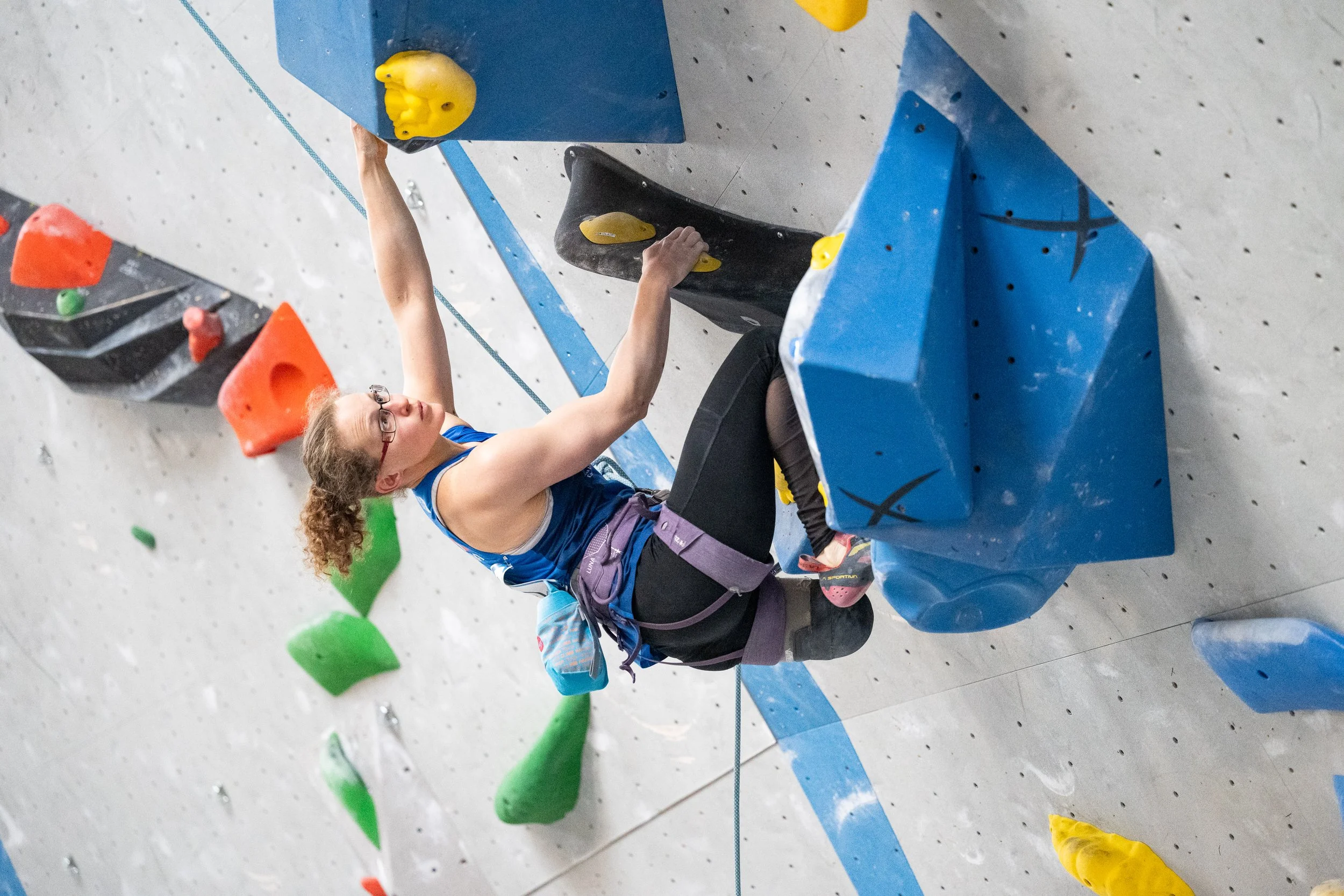 Une femme fait de l'escalade en salle, portant un harnais et regardant vers le haut, avec des prises de couleur verte, orange, jaune et bleue sur le mur d'escalade.