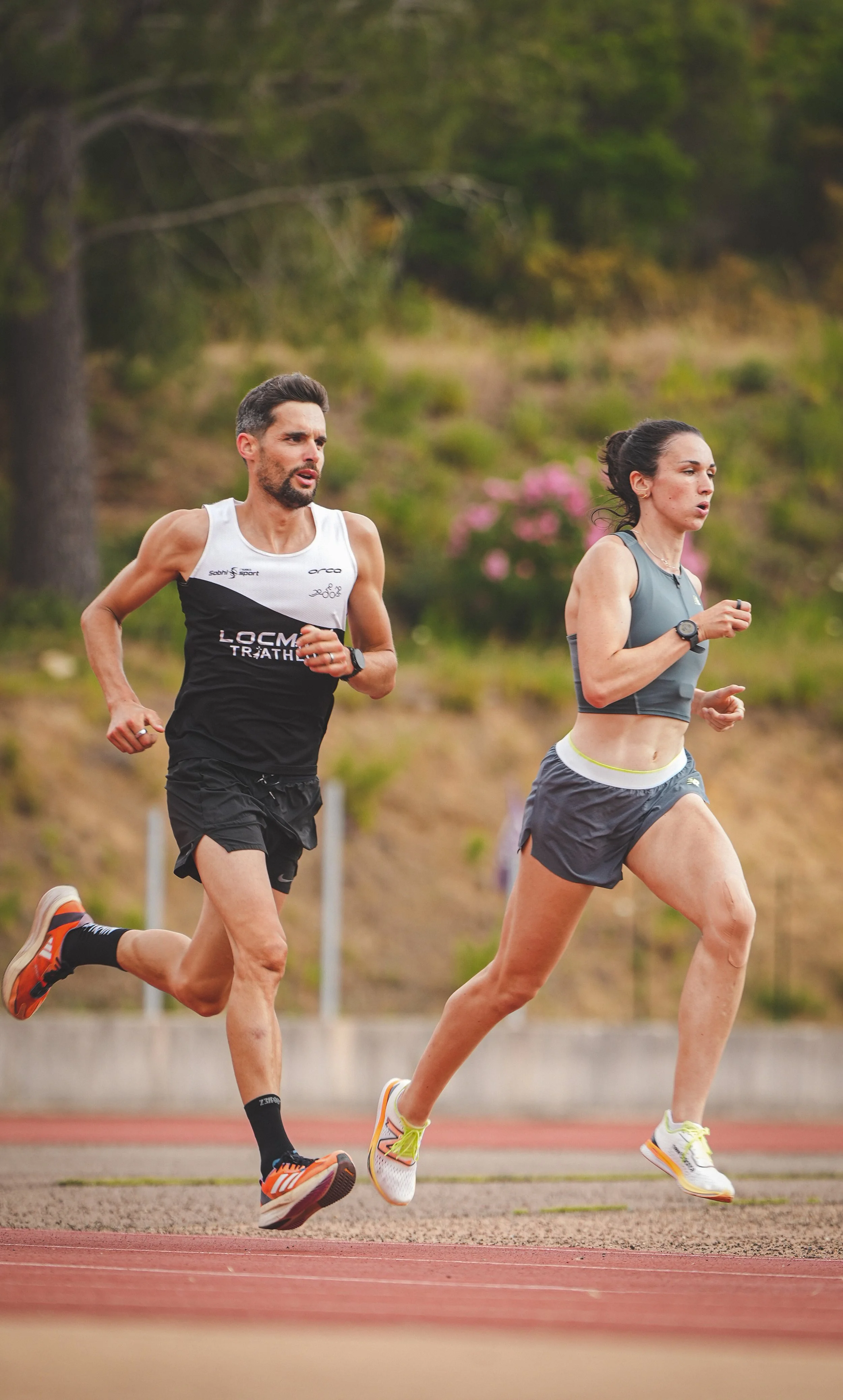 Deux personnes courent sur une piste d'athlétisme en plein air, un homme avec un débardeur noir et blanc et une femme avec un débardeur gris et un short noir, entourés d'arbres et de végétation.