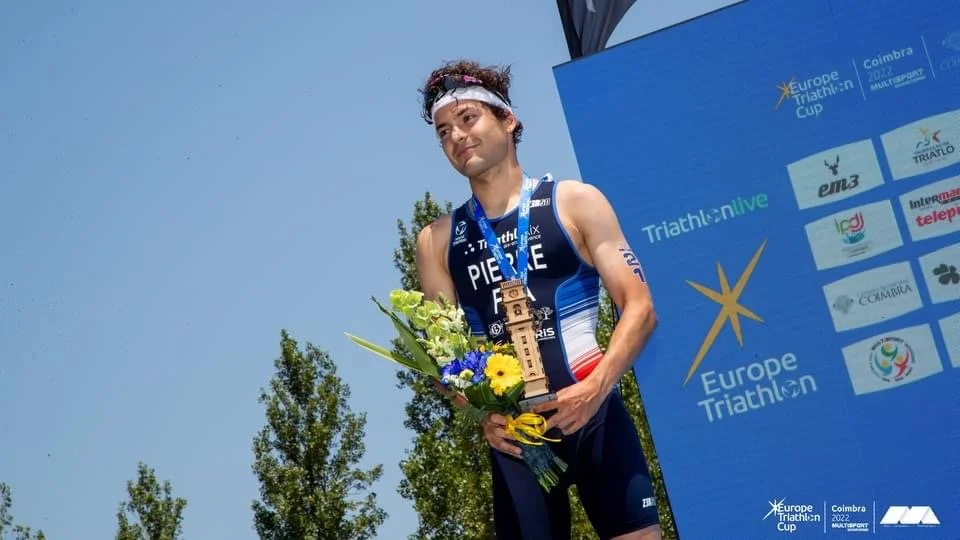 Triathlète masculin reçoit une médaille d'or lors du triathlon Europe Cup à Coimbra, tenant une bouquet de fleurs, portant un costume bleu avec le drapeau français, arrière-plan de panneau d'événements et ciel clair.