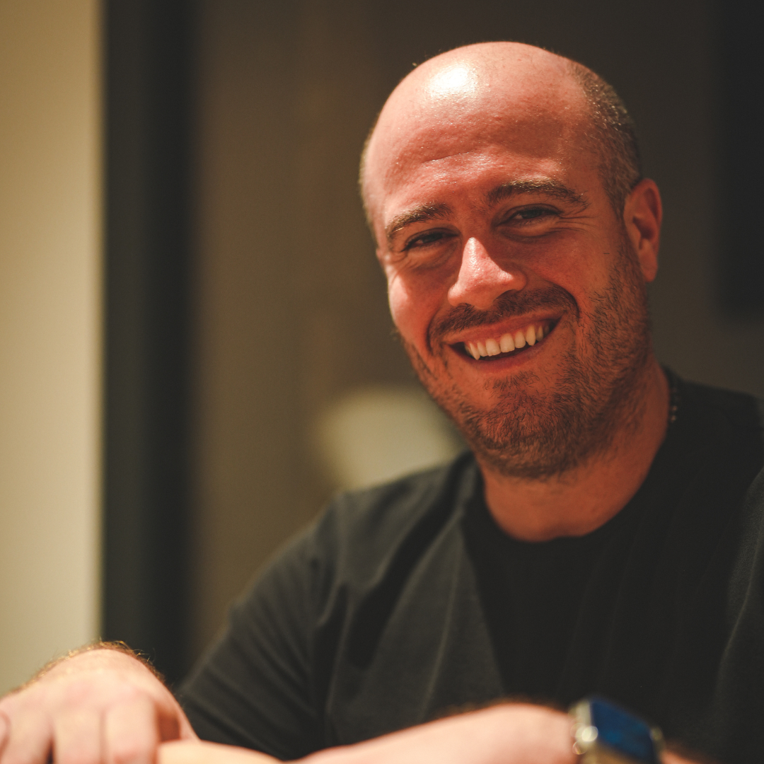 Homme souriant avec une barbe, portant un t-shirt noir, assis en intérieur.