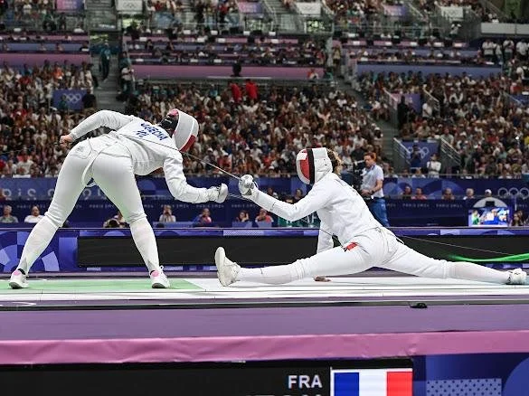 Auriane Mallo-Breton en compétition aux Jeux Olympiques, engagés dans un duel, portant des tenues blanches et des masques protecteurs, avec un public en arrière-plan.