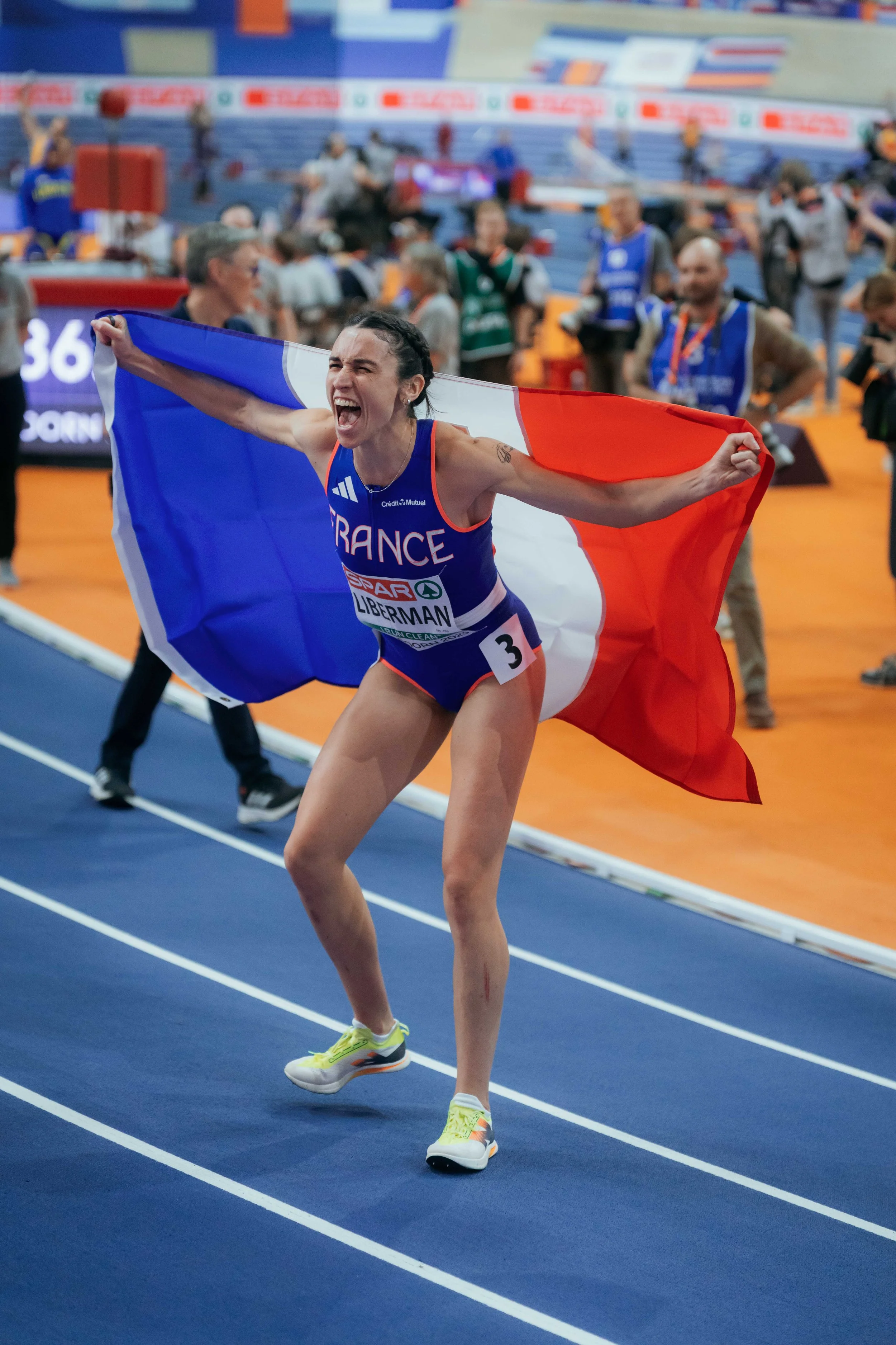 Une athlète française célèbre sa victoire en tenant un drapeau français lors d'une compétition d'athlétisme en intérieur.