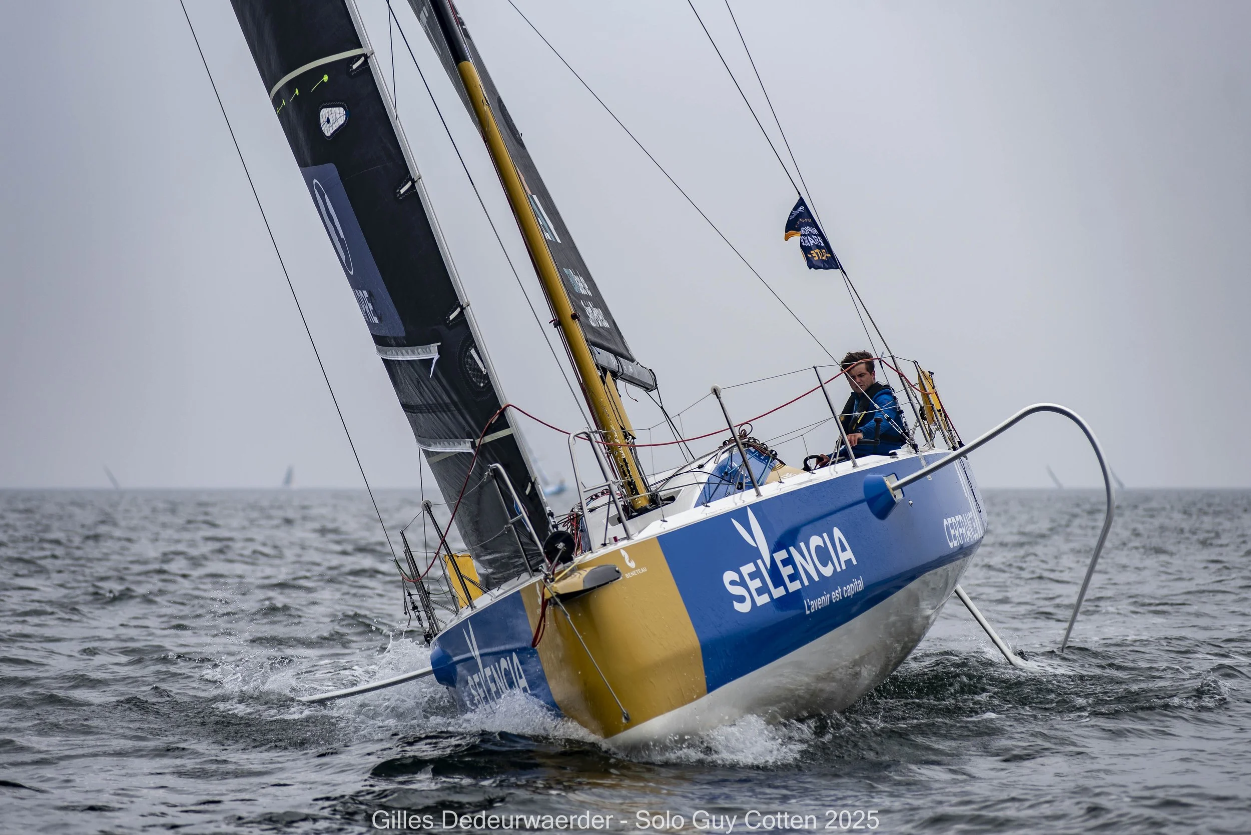 Un voilier bleu et jaune portant le nom 'SELENCIA' naviguant sur la mer par temps nuageux, avec un seul marin à bord. La voile est en partie déployée et l’eau est agitée.