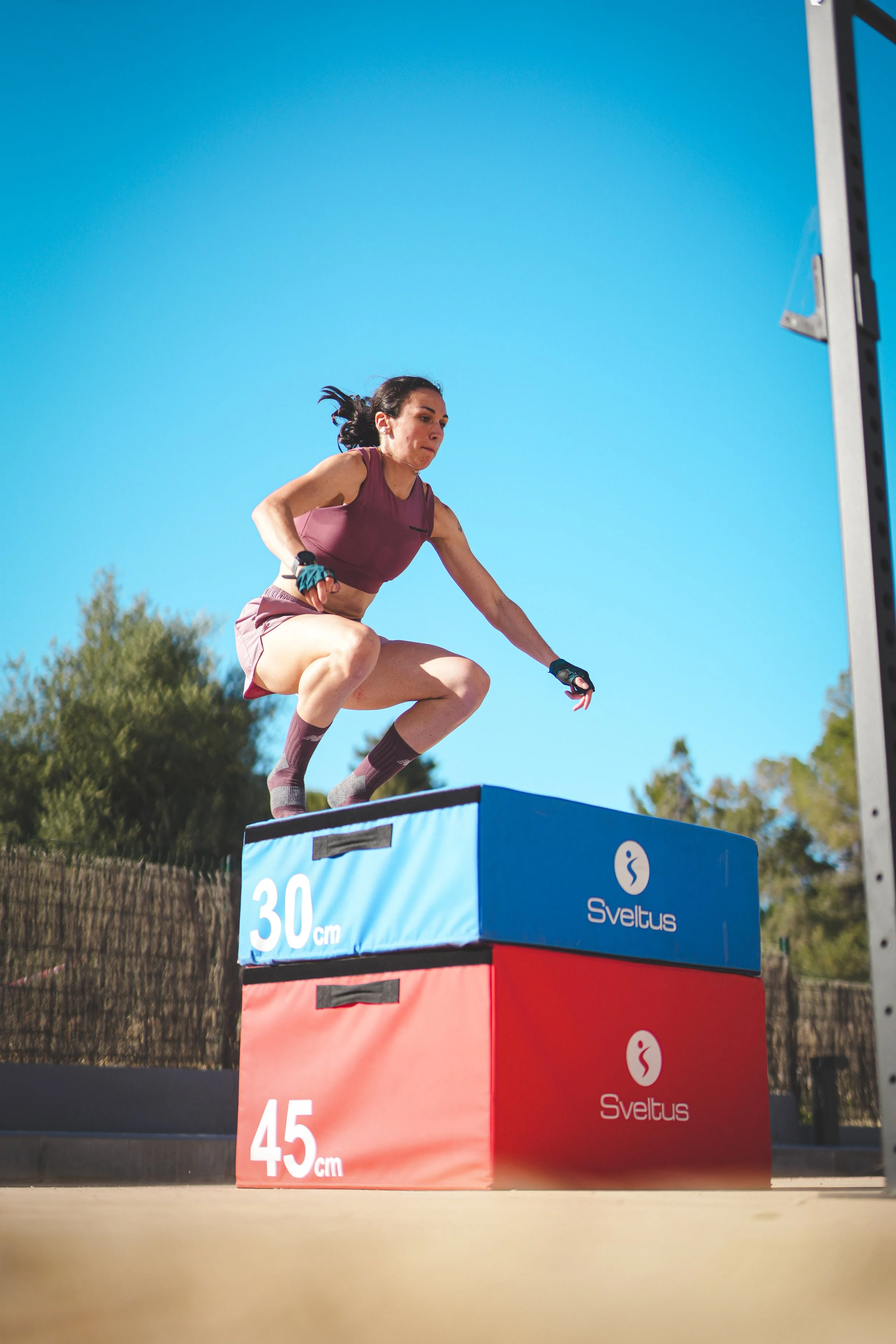 Une femme en tenue de sport saute sur des bancs de saut, à l'extérieur par une journée ensoleillée, avec un ciel clair et des arbres en arrière-plan.