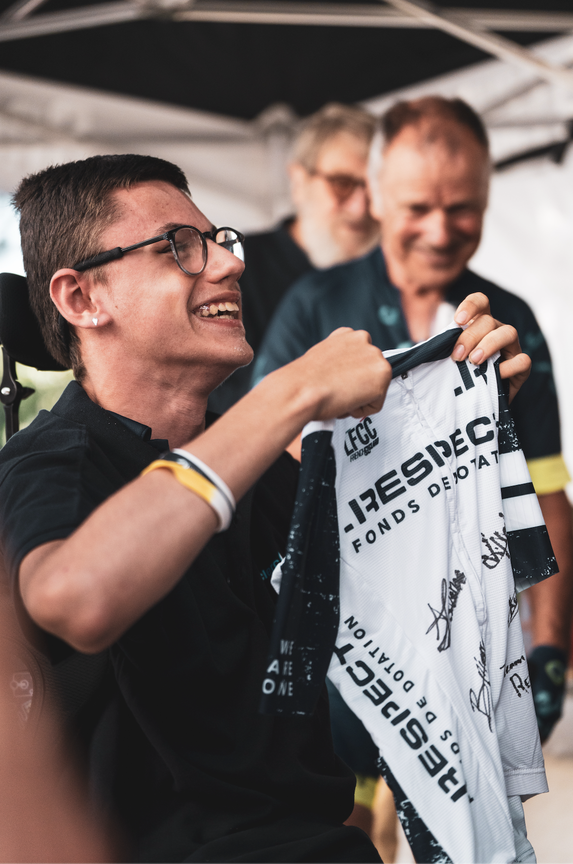 Une personne souriante tenant un maillot de cyclisme signé, entourée de deux autres personnes en arrière-plan, lors d'un événement en plein air.