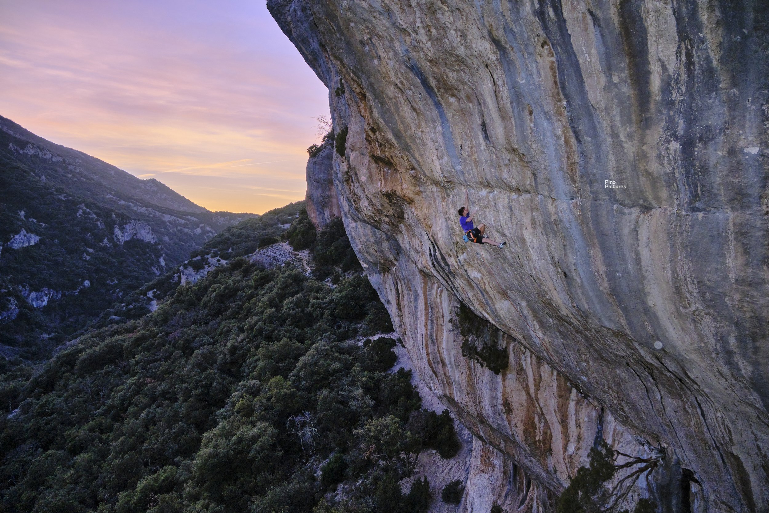 Cramponnage sur une falaise rocheuse au coucher du soleil, avec des montagnes en arrière-plan et un ciel coloré.