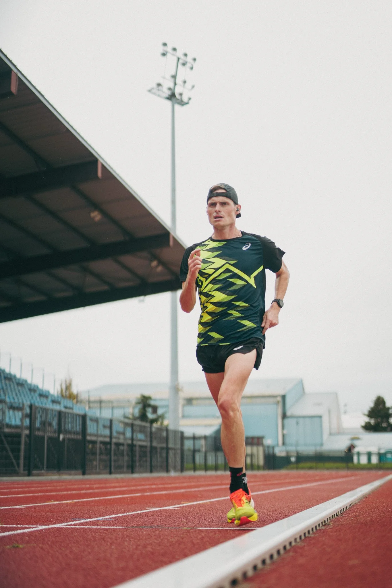 Aurélien Quinion court sur une piste d'athlétisme en extérieur, portant un t-shirt noir avec des motifs jaunes fluo, un short noir, des chaussures de course néon, un sweat noir attaché autour de la taille, et une montre