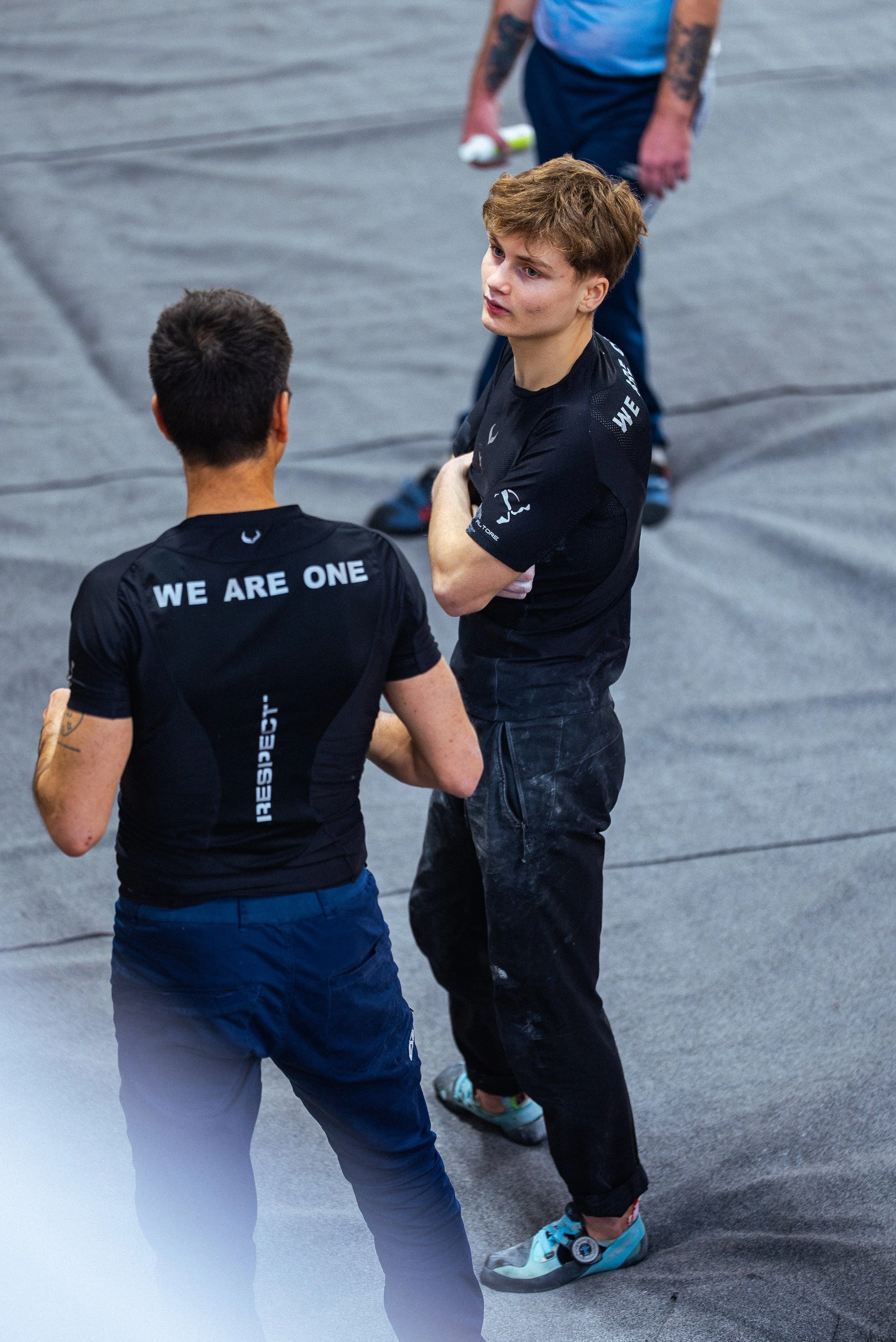 Deux jeunes hommes discutent, portant des tenues de sport noires avec des inscriptions telles que "WE ARE ONE" et "RESPECT". Ils se tiennent dans un espace intérieur avec un sol en toile.