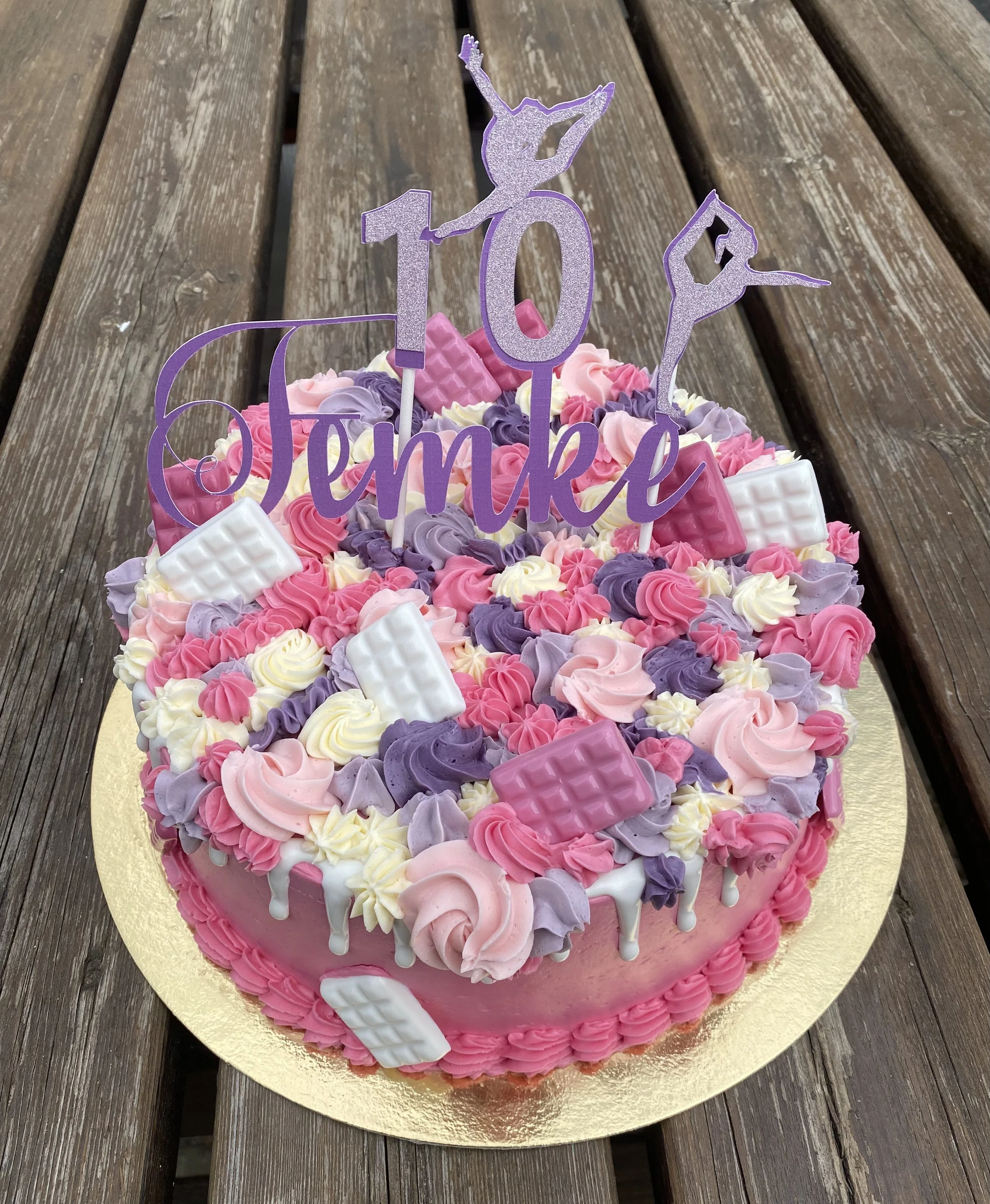 Een roze en paarse verjaardagstaart met decoraties en een '10' cijfer topper, op een gouden taartonderlegger op een houten tafel.