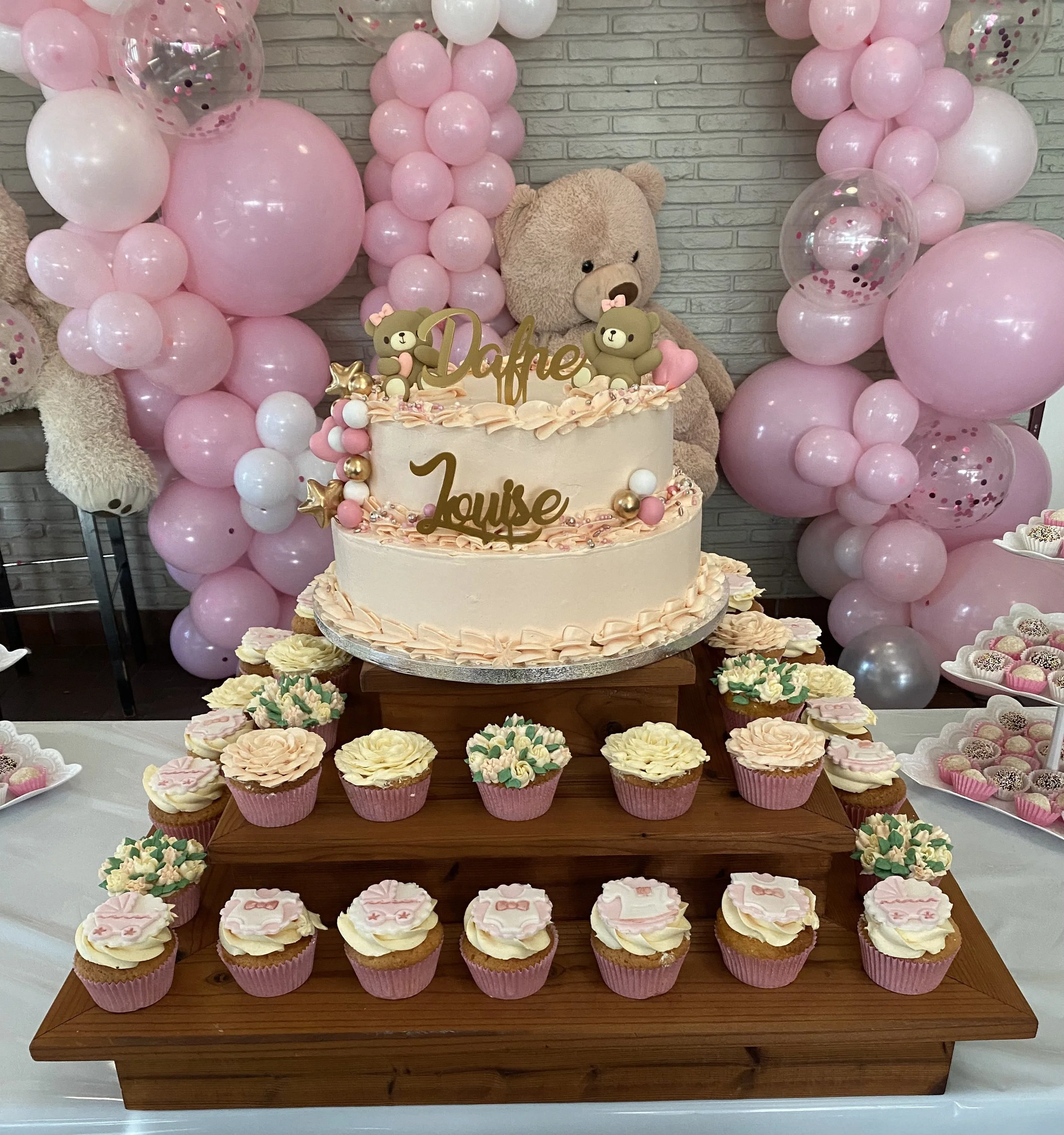 Een feesttafel met een roze en witte taart en cupcakes, versierd met teddyberen en ballonnen in roze en wit, voor een babyfeest of babyshower.
