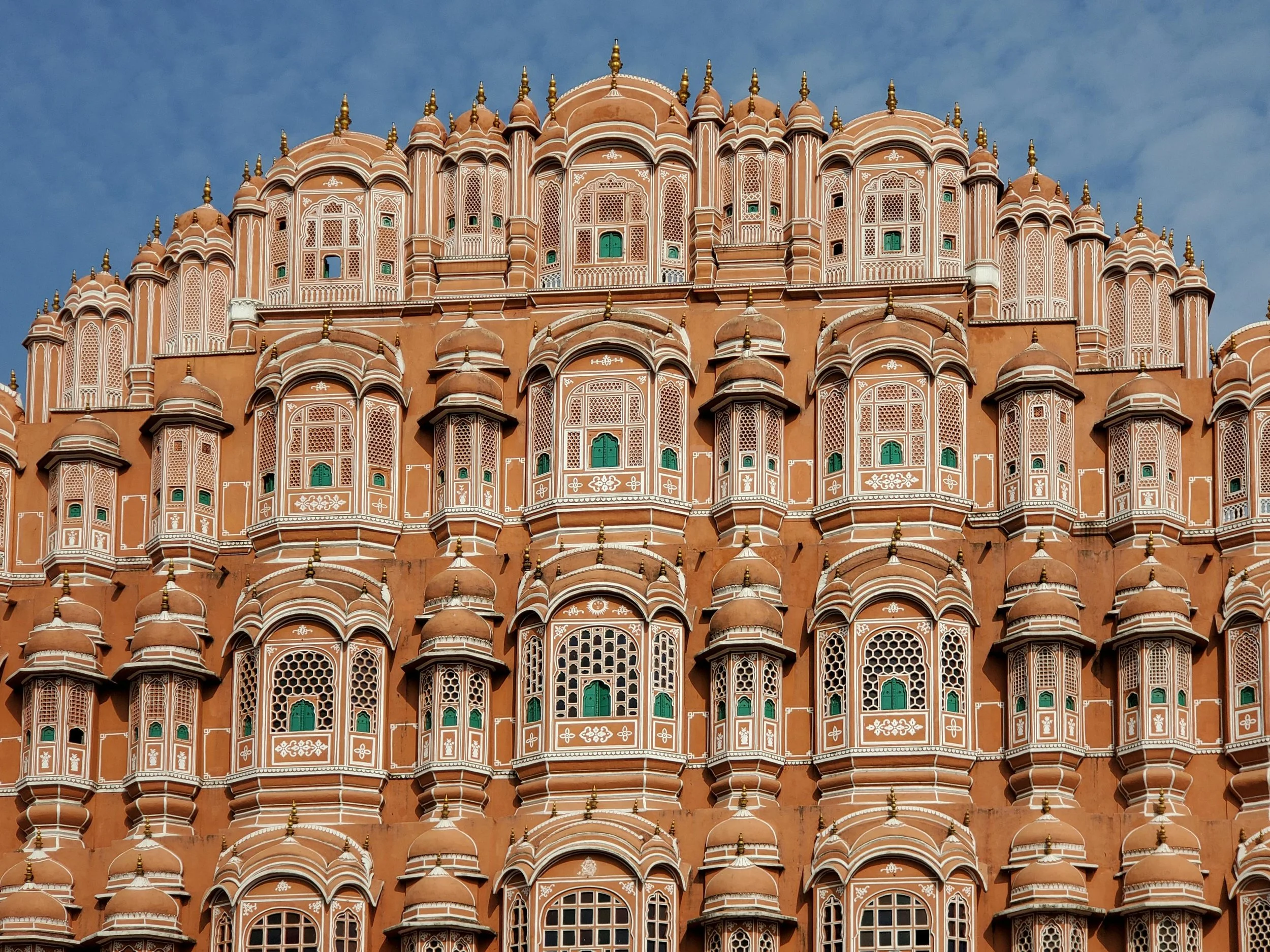 The façade of the Hawa Mahal in Jaipur, India, featuring intricate lattice windows and small balconies in a pink sandstone structure against a blue sky.