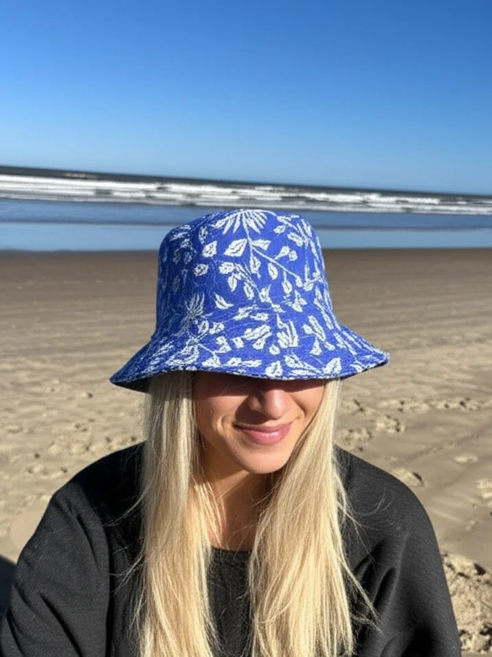 Young woman with long blonde hair wearing a blue and white patterned sun hat, sitting on a sandy beach with the ocean and waves in the background under a clear blue sky.