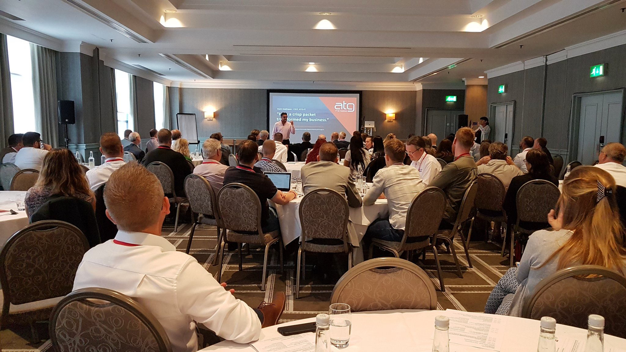 A man in a white shirt speaking at a technology conference, with an audience of business professionals seated in a conference room. Behind him is a banner with a group's logo and the words "Network Up" and "The Power of Technology Community." A projector screen displays a presentation on the left.