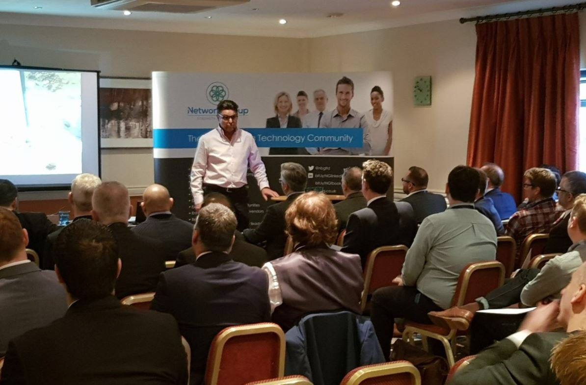 A man in a white shirt giving a presentation to an audience seated in a conference room. There is a large banner behind him with images of diverse people and the text 'Network Group' and 'The UK Technology Community.' A projector screen is visible on the left side of the room.