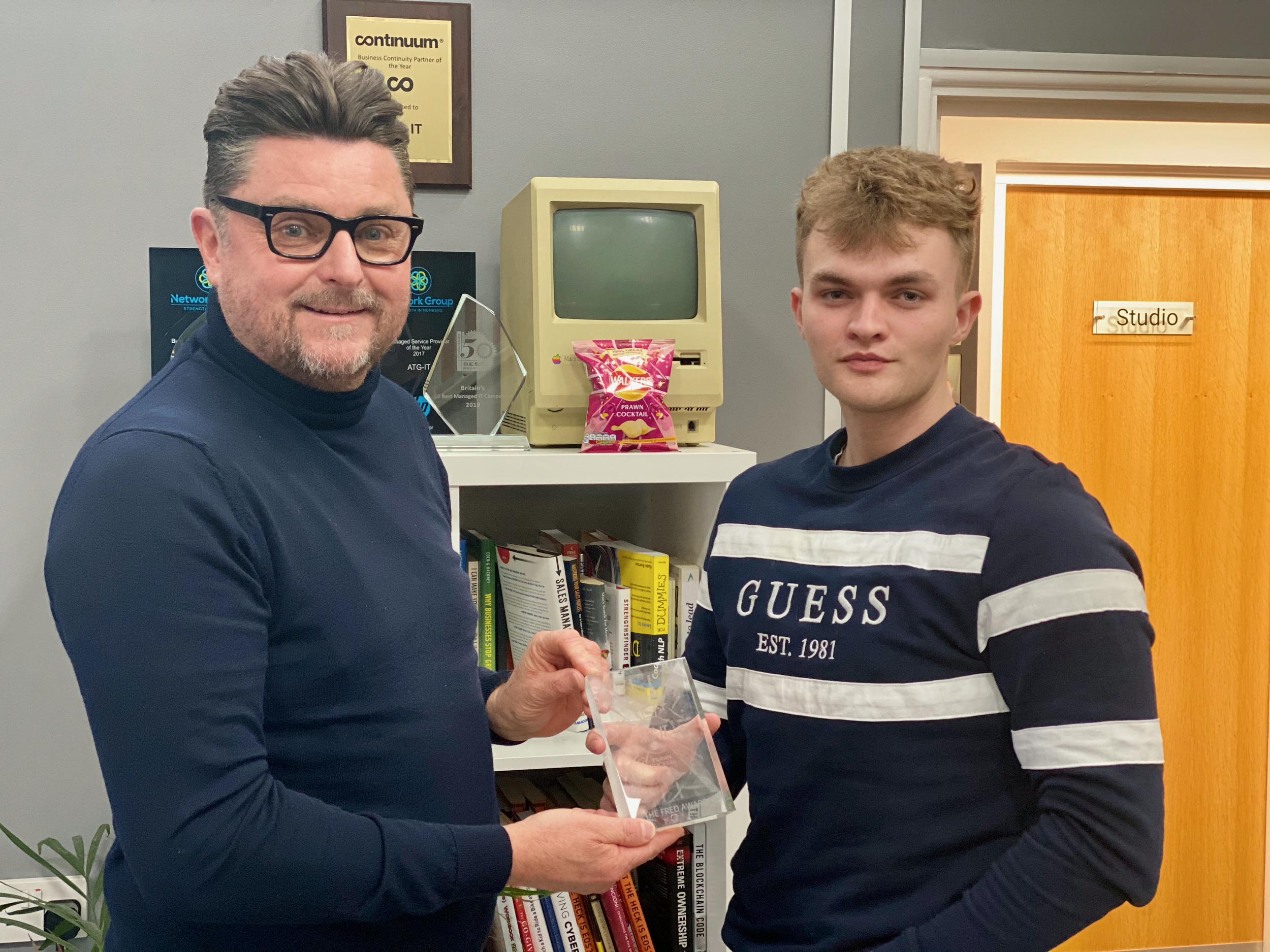 A man presenting at a technology event with an audience seated in front of him. There is a projection screen and a banner with people’s images and technology-related text behind him.