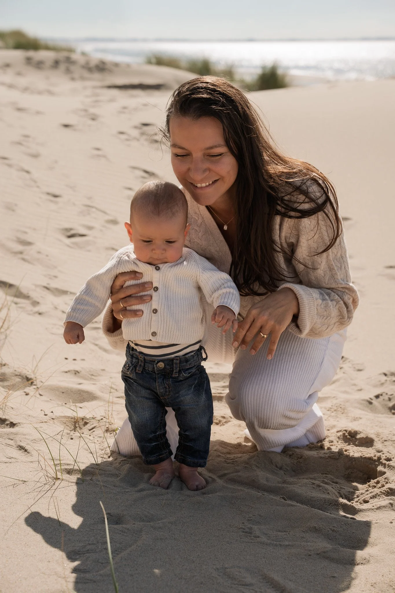 A woman and a baby on a sandy beach during daytime, with the woman kneeling and holding the baby, both looking down at the sand, with the ocean in the background.