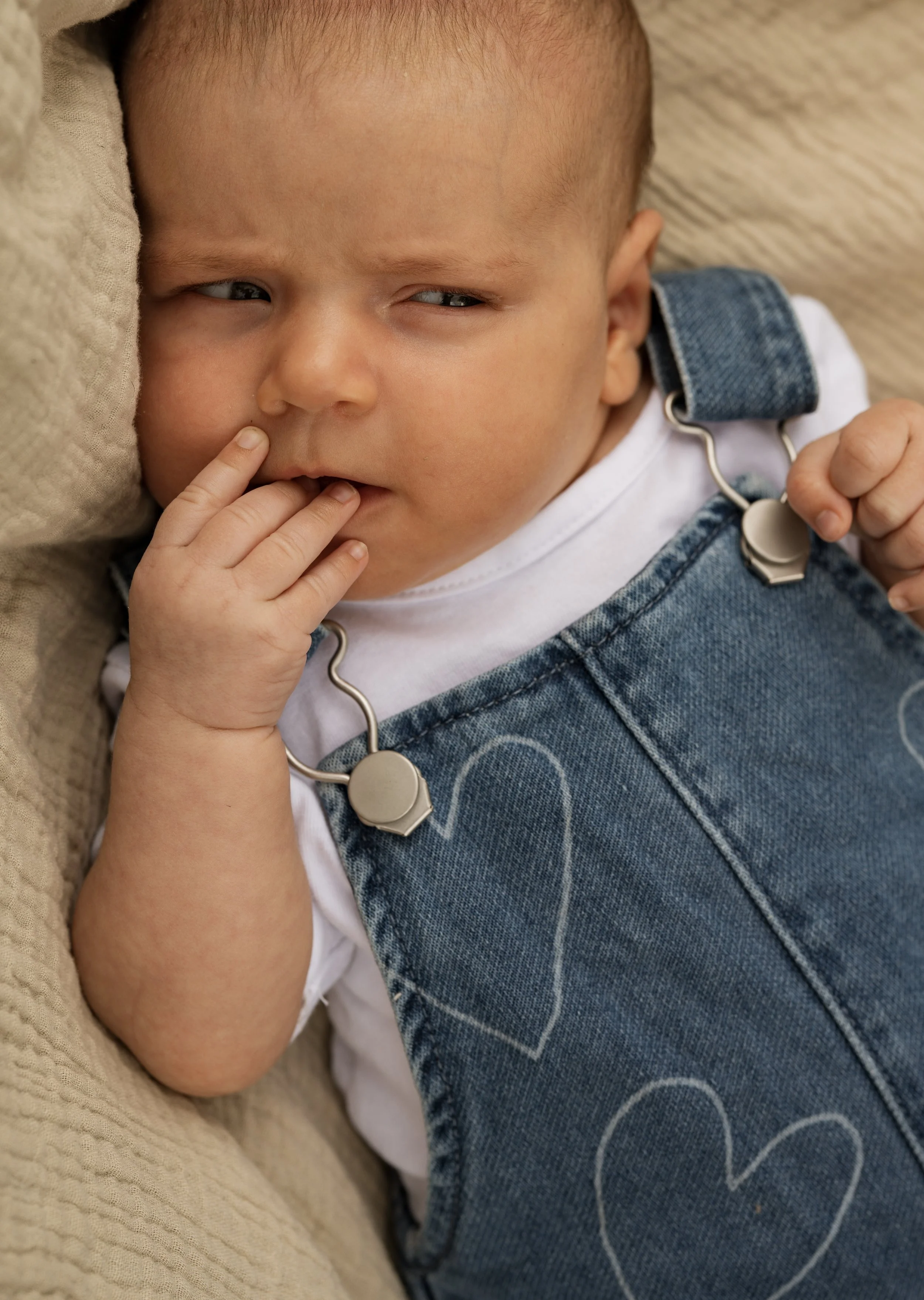 Close-up of a baby lying on a beige blanket, wearing denim overalls with hand-drawn white hearts and a white shirt, with a curious expression, finger near their mouth, and a pacifier clip attached to the overalls.