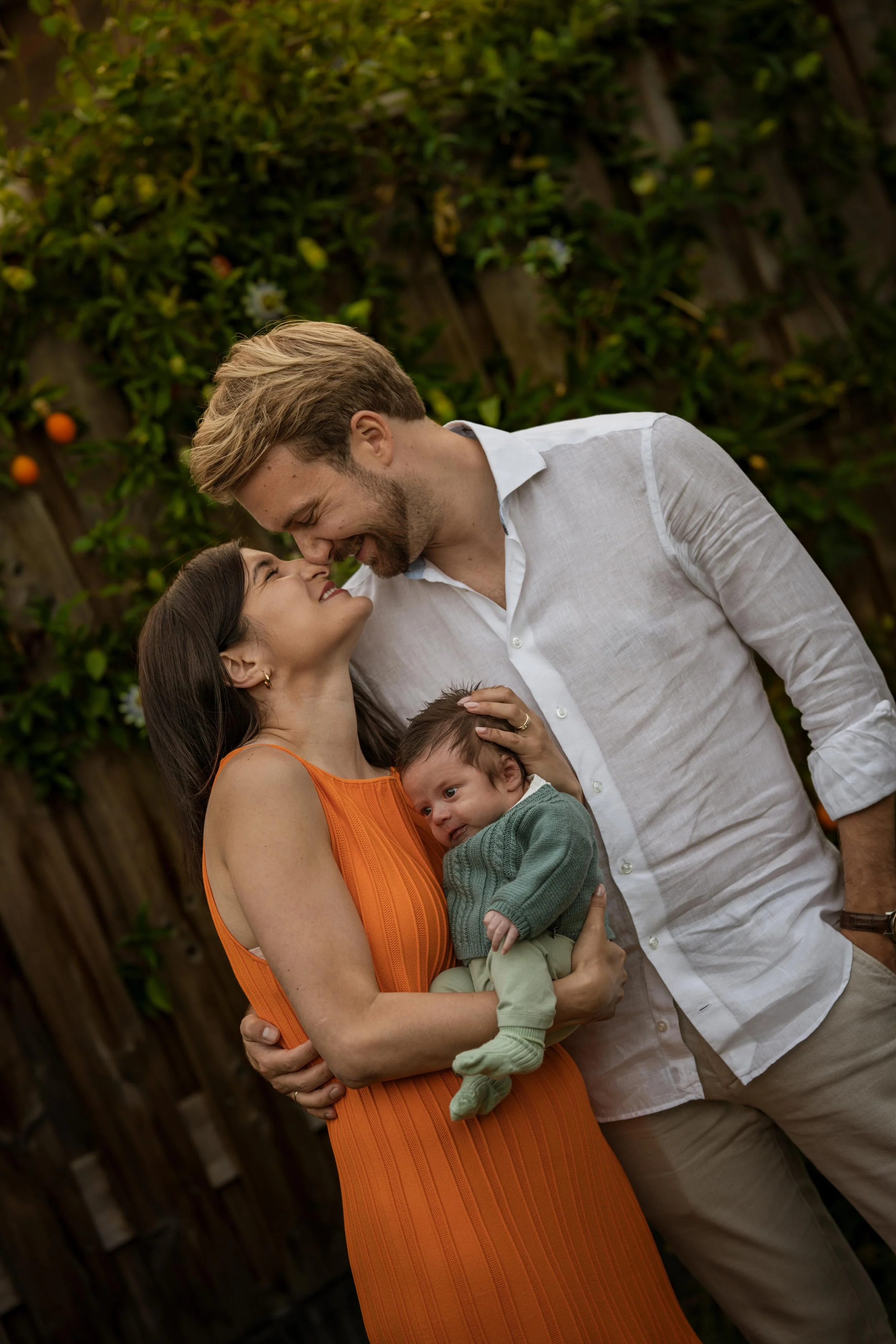 A family of three, a woman in an orange dress, a man in a white shirt, and a baby in green, sharing a joyful moment outdoors.