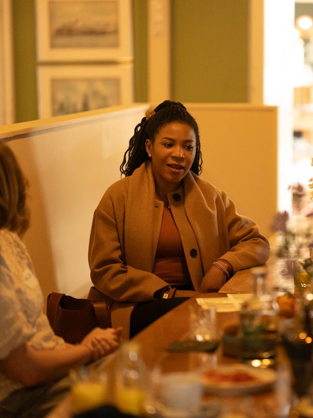 Women sitting at a dinner table with food and drinks, engaging in conversation in a warmly lit room. Event photography