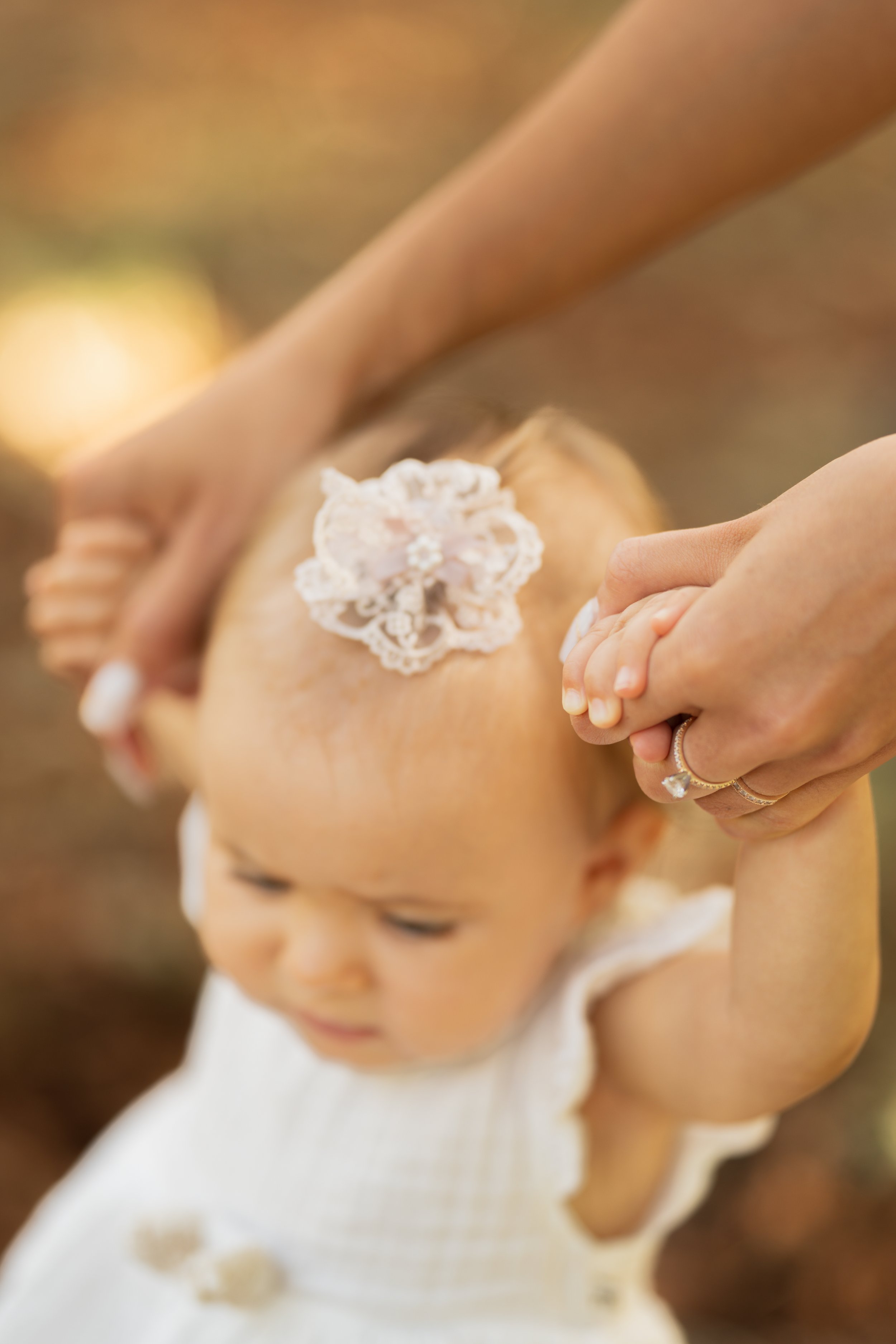 A baby girl holding an adult's hand outdoors, with lace decoration on her head and a white dress.