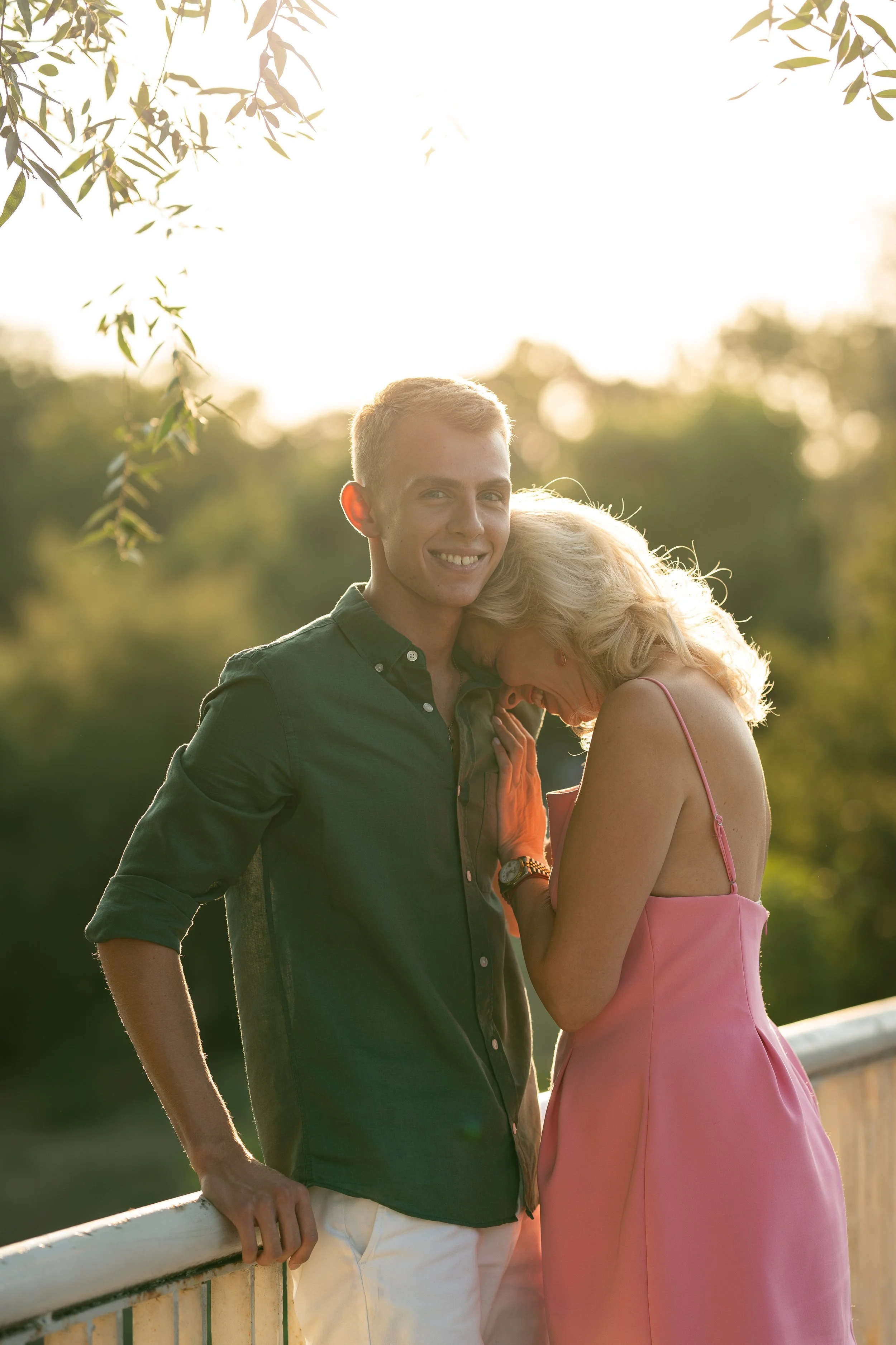 A young man and an older woman share a laugh outdoors during sunset, standing by a white railing with trees in the background.