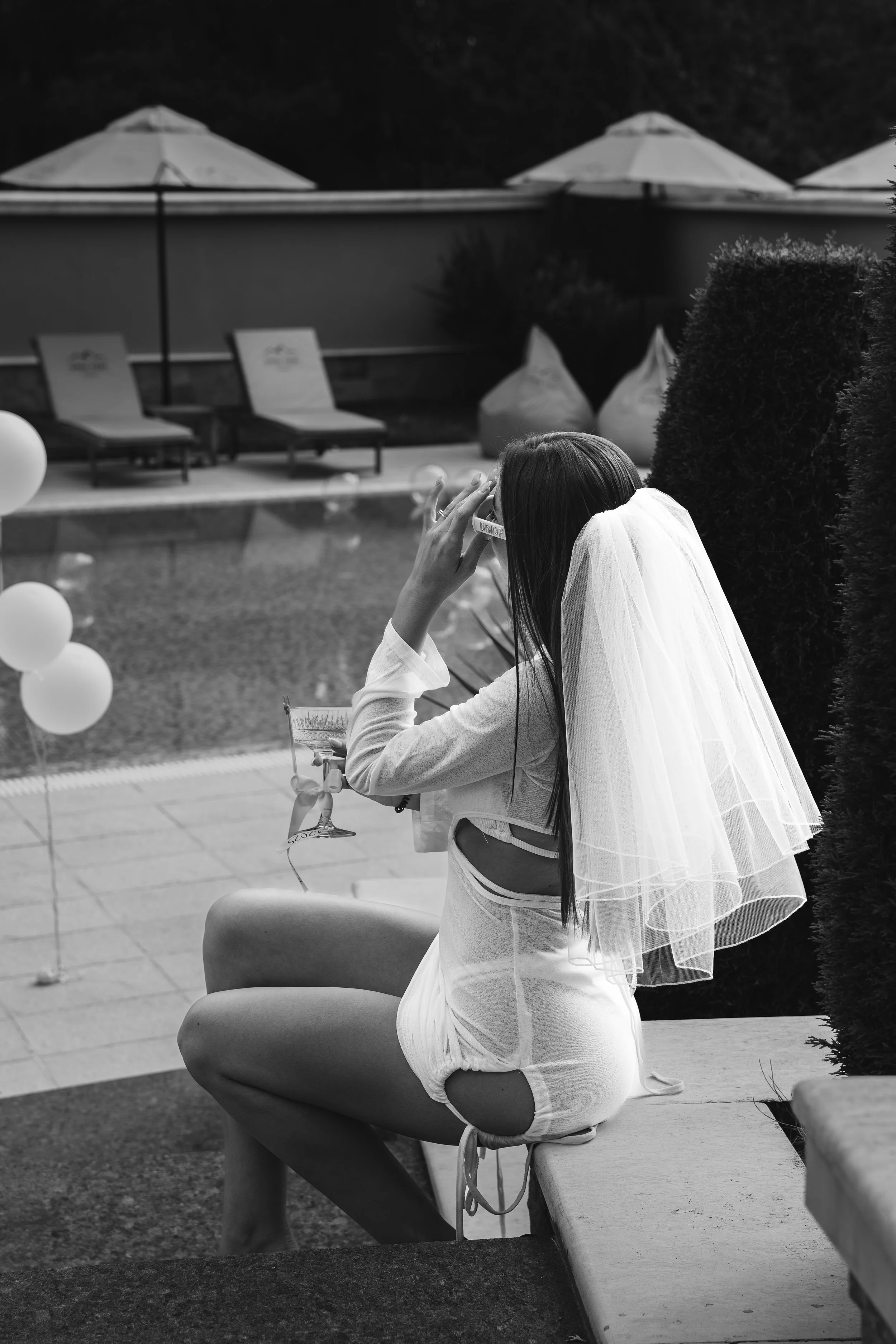 A woman wearing a bridal veil, shorts, and a long-sleeve top sitting outdoors near a swimming pool, holding a drink and sunglasses, with balloons and poolside chairs in the background.