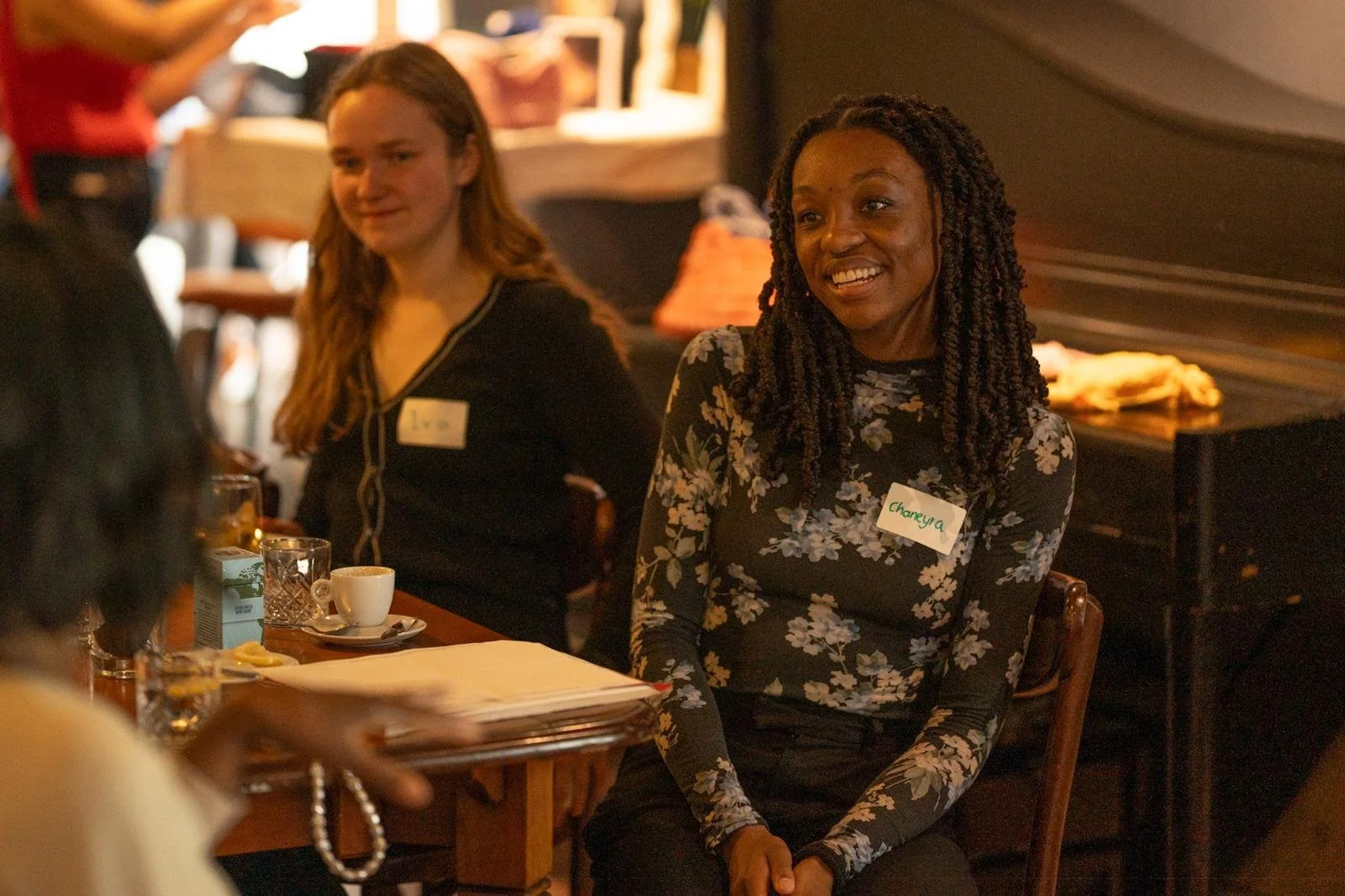 Two women sitting at a table in a cozy restaurant or cafe, smiling and engaging in conversation. They have name tags on, one with the name "Chareya" and the other with a name partially visible.