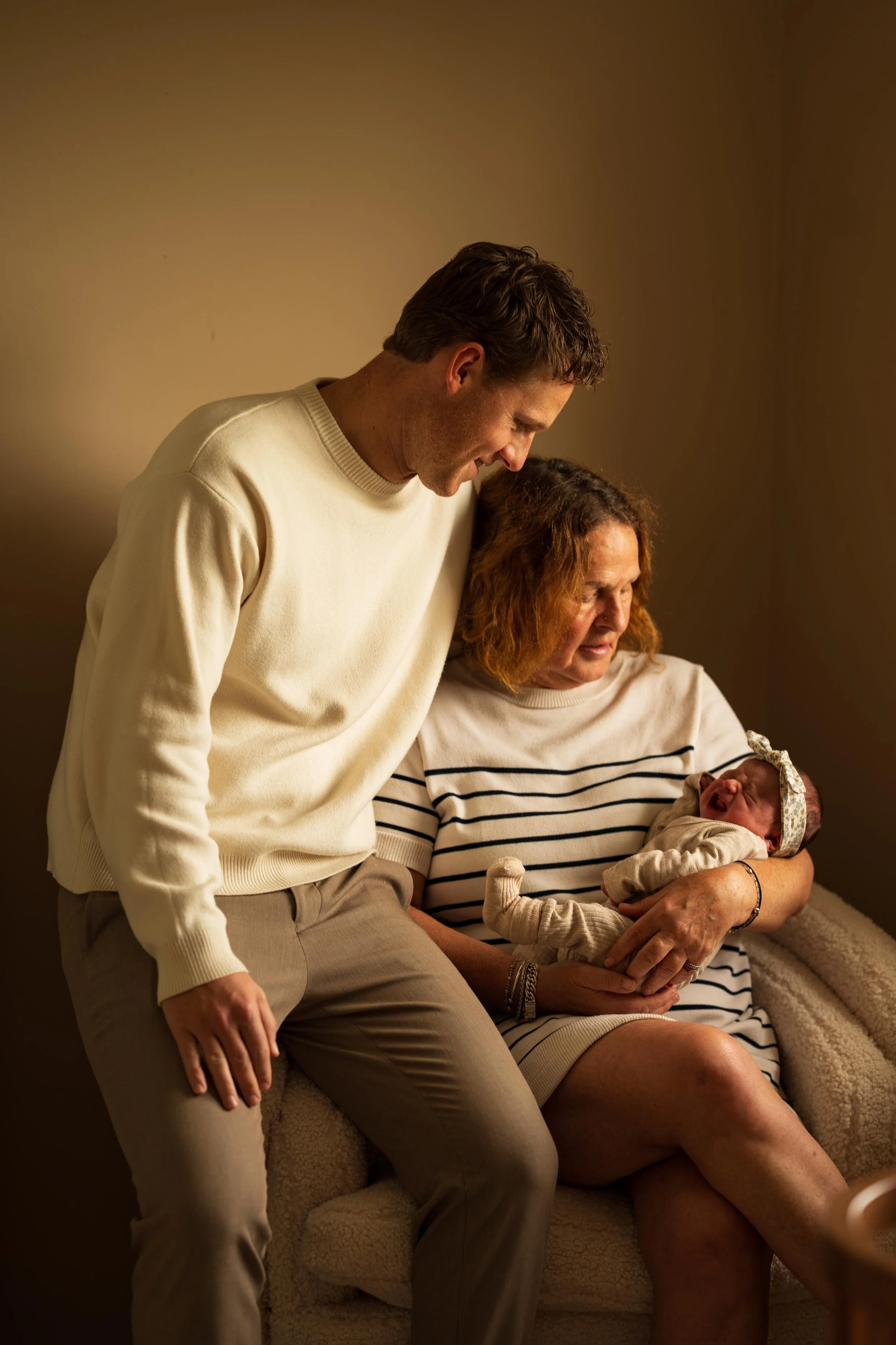 A young man and an older woman watch a newborn baby. The woman is sitting on a chair, holding the baby, and the man is leaning over her, smiling. The scene is warmly lit and appears to be in a cozy indoor setting.