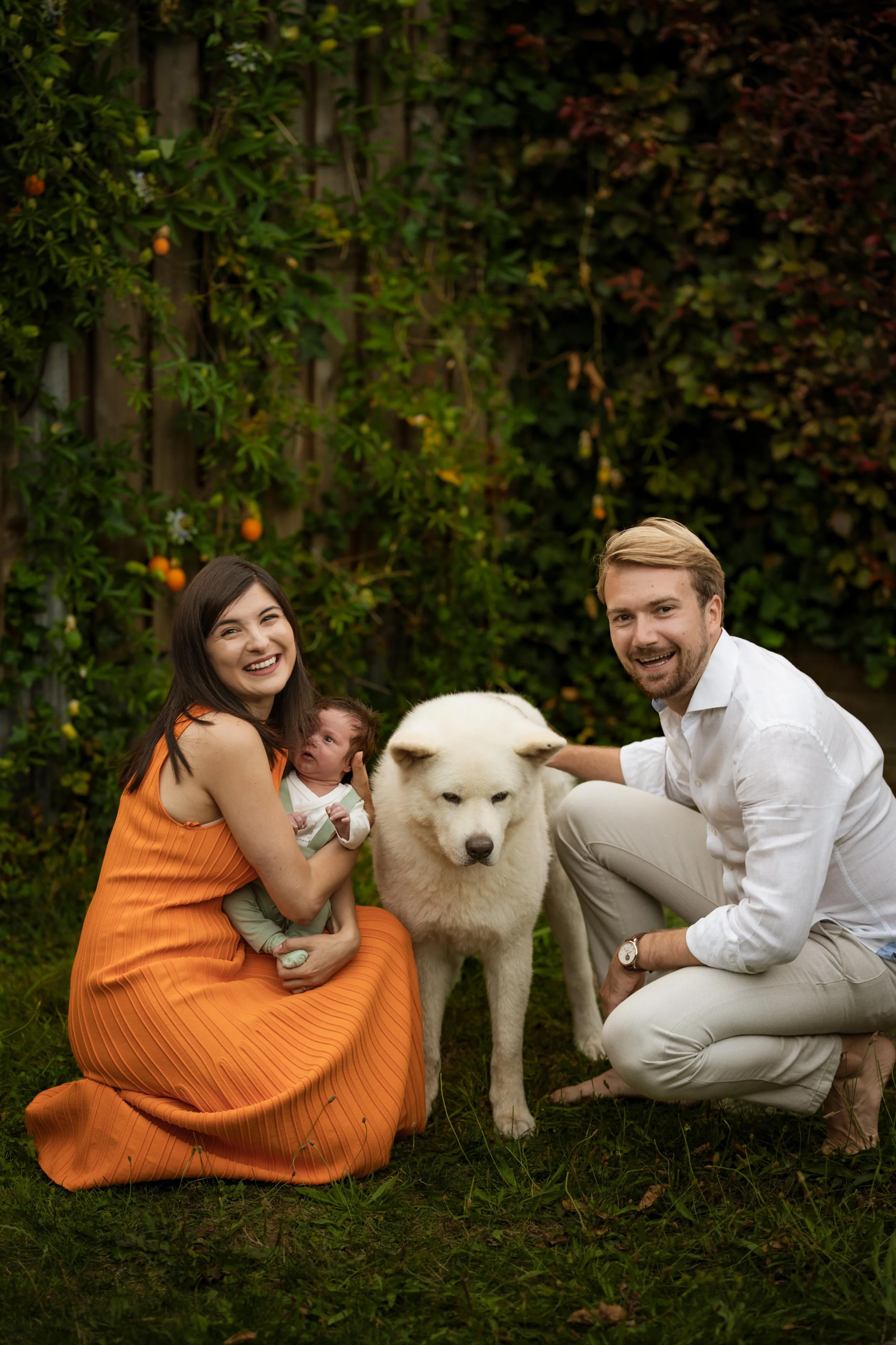 A smiling family of three, including a woman in an orange dress, a man in a white shirt, a newborn baby, and a large white dog, gathered in a green garden with fruit-bearing plants.