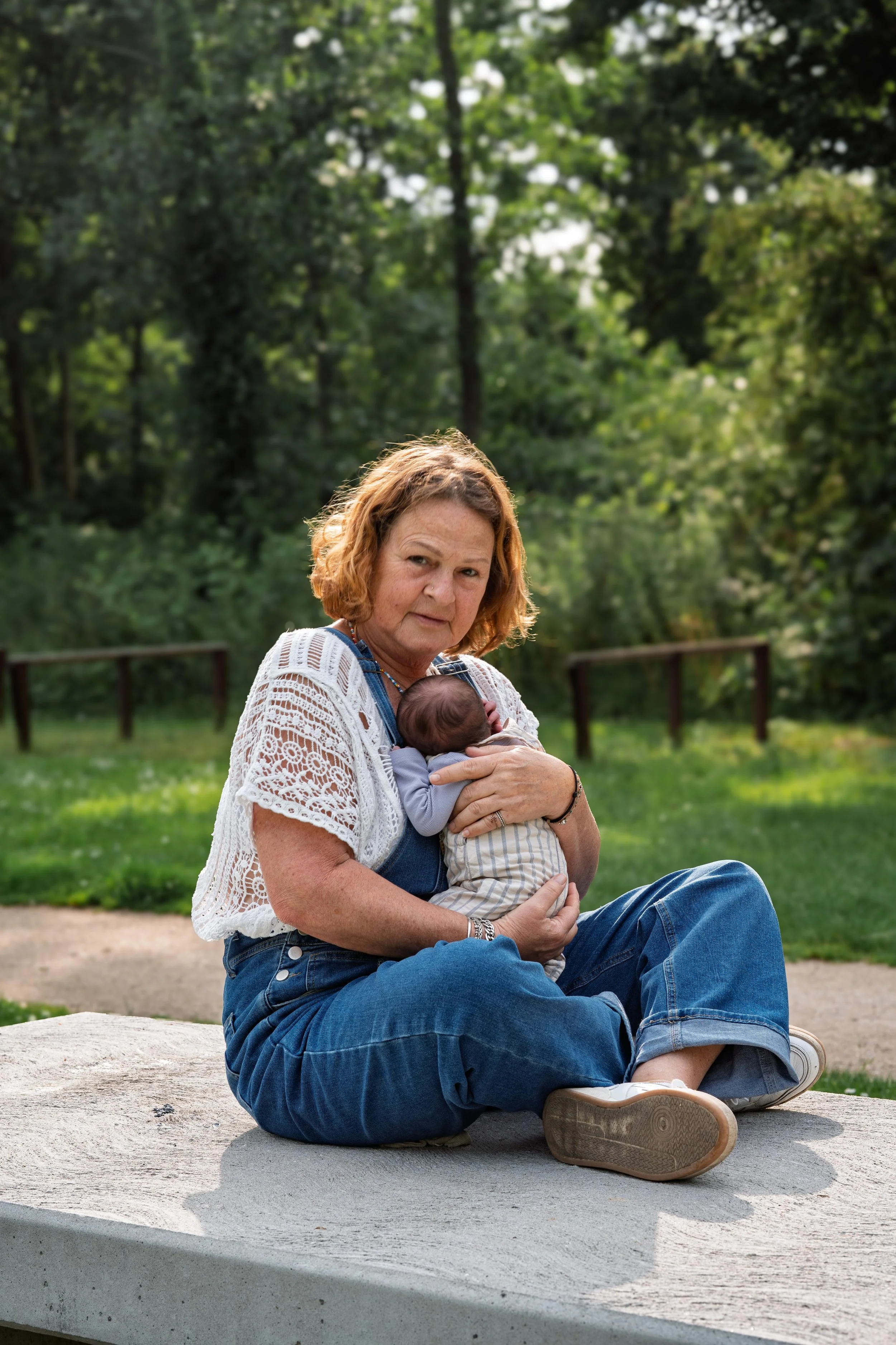 An older woman sitting outdoors on a concrete ledge, holding a sleeping baby in her arms, with a background of green trees and grass.