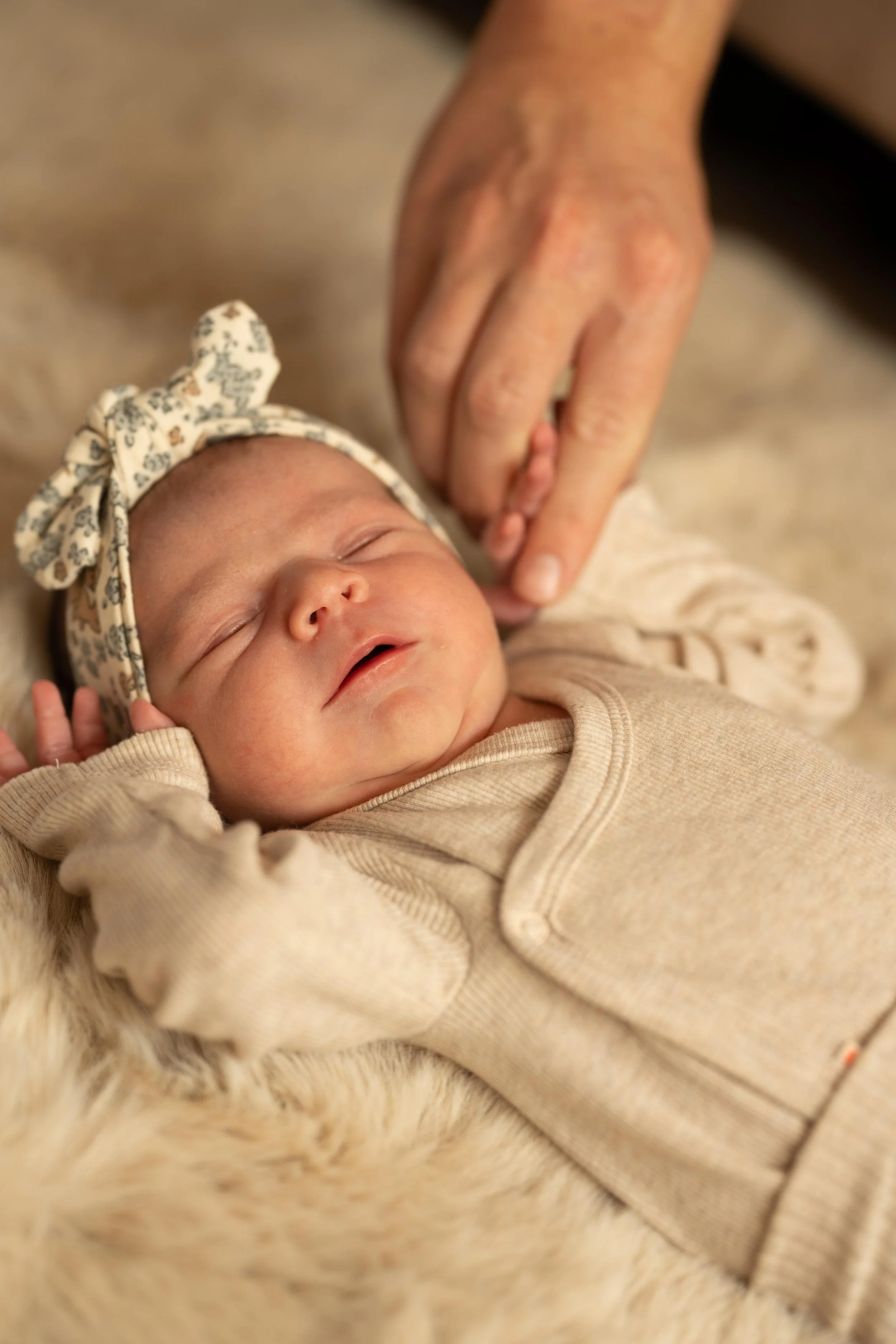 A sleeping baby girl wearing a beige outfit and a floral headband, resting on a fuzzy blanket, while an adult's hand gently touches her head.