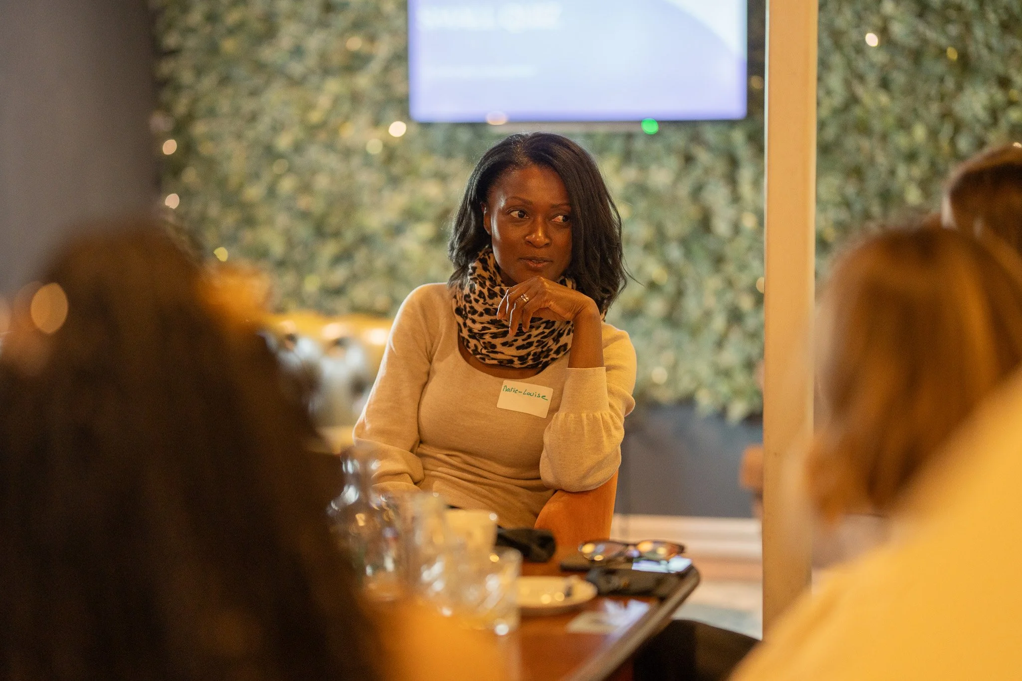 A woman with dark hair and a leopard print scarf sitting at a table with people, in a room with a large screen and a green plant wall in the background.