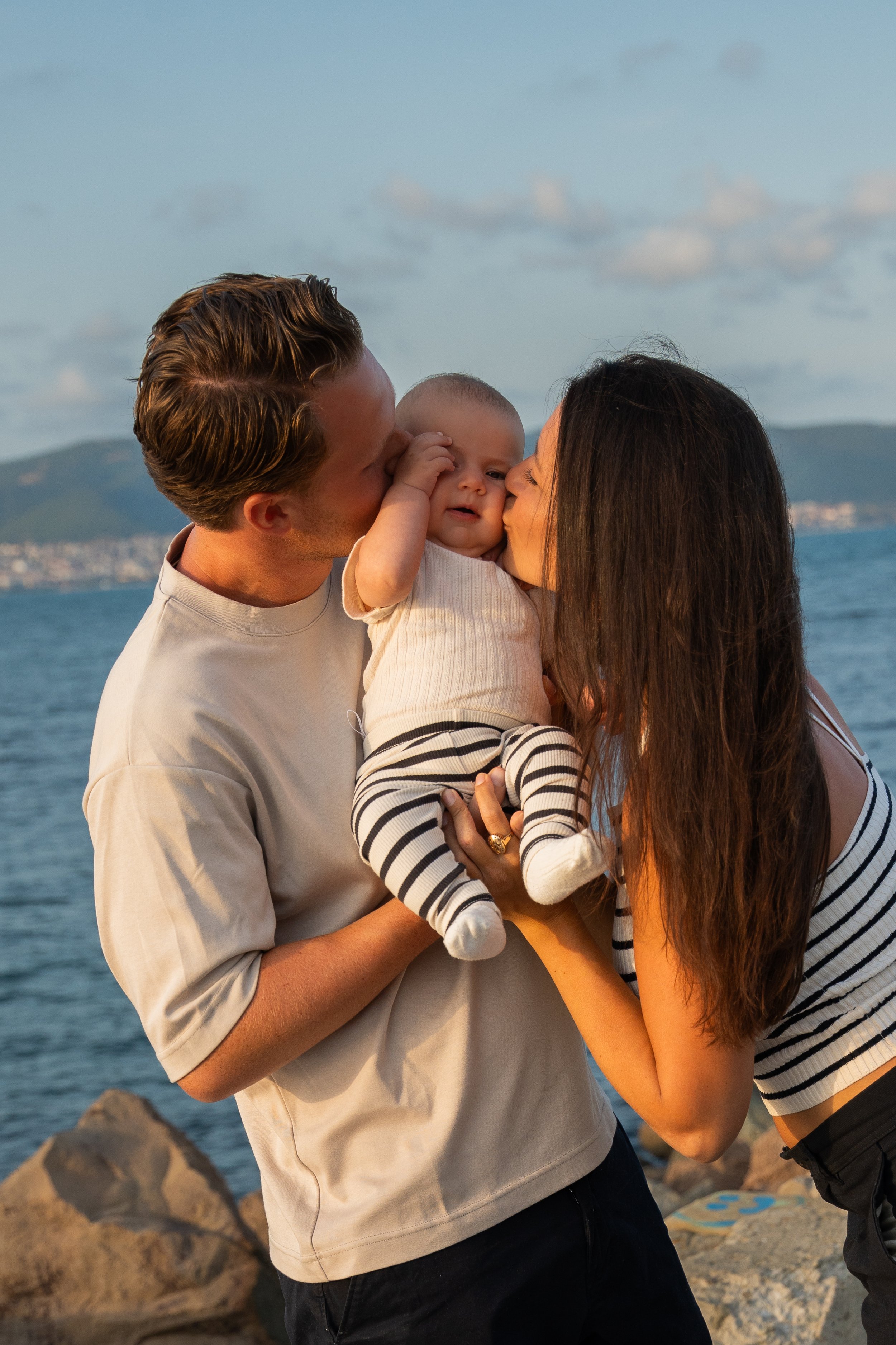 A young family of three at the beach, with a man and woman kissing their baby while holding him up, with a scenic ocean and mountains in the background.